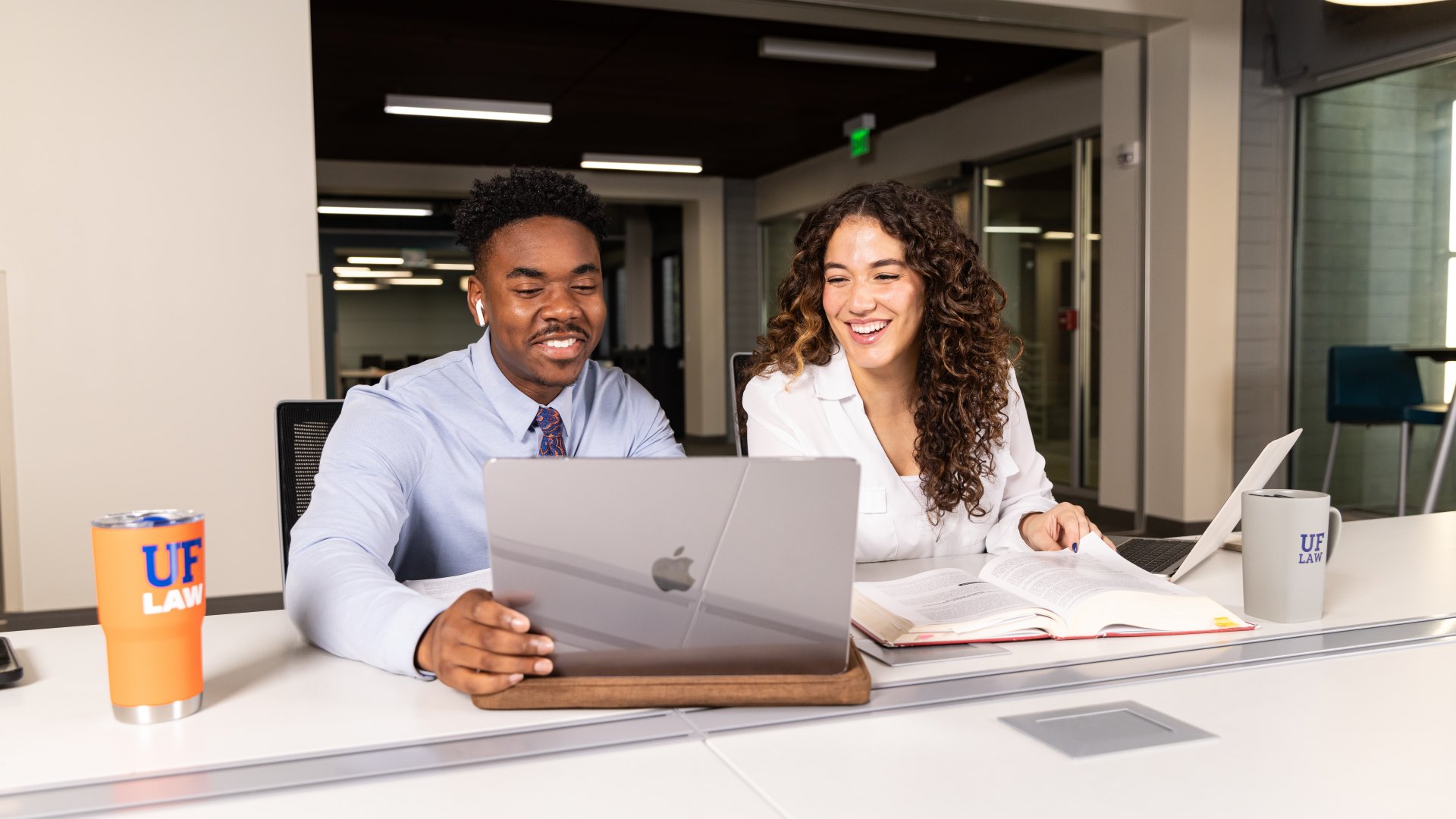 Two students in professional attire sit at a table looking and smiling at a laptop