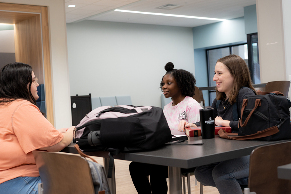 Three students sit at a table, socializing, in the Commons