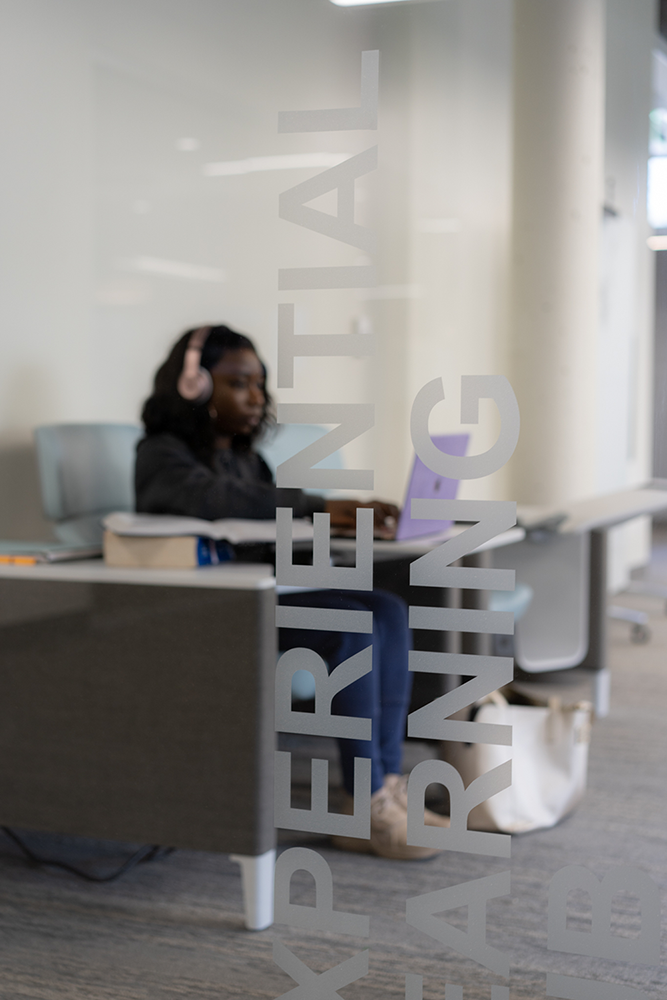A student works on a laptop wearing headphones, seen through glass with Experiential Learning etched on the surface