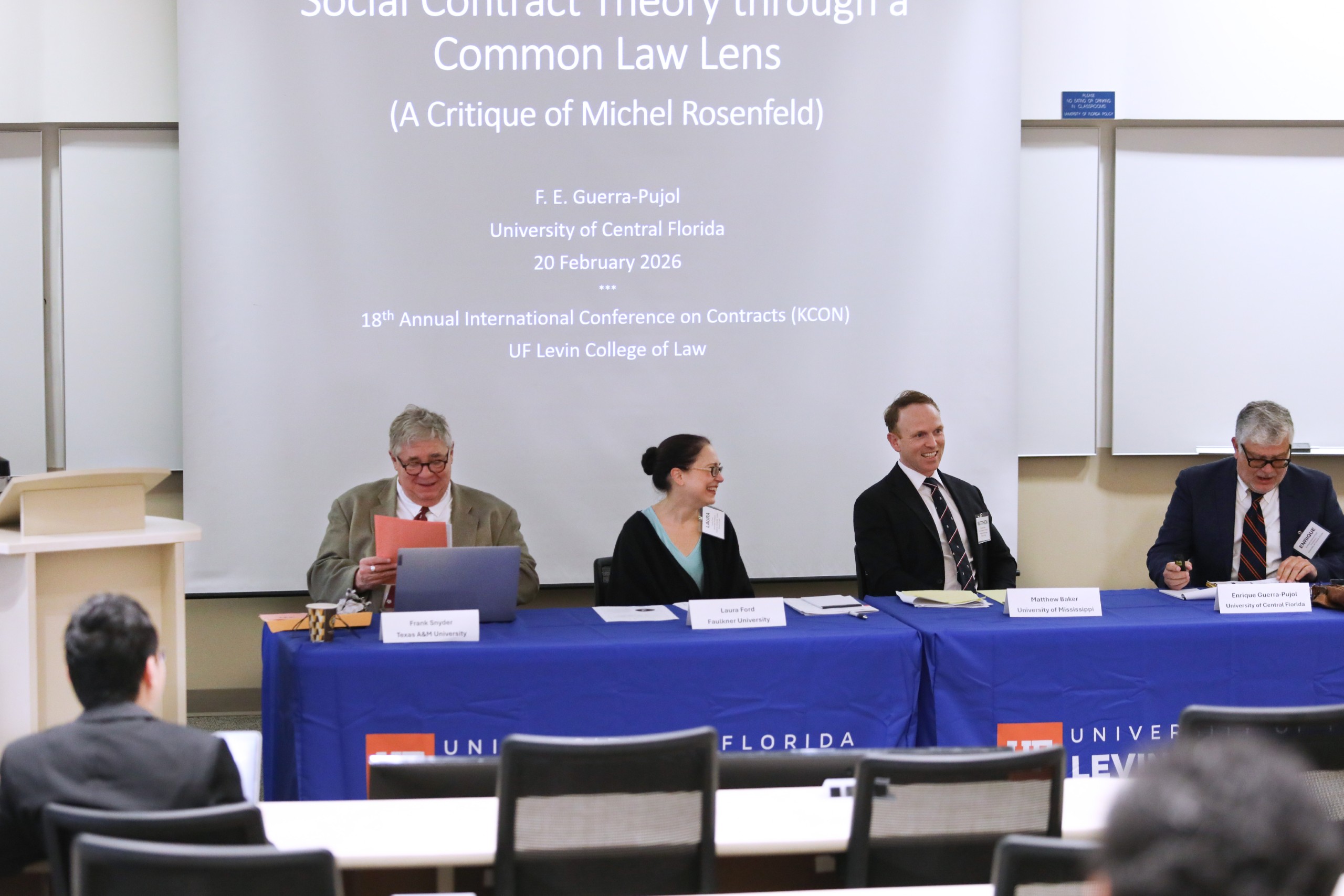 Four people sit at a long table in a lecture room facing the audience laughing as they talk to each other and to the audience (not pictured). Behind them is a slide projection that reads "Social Contract Theory Through a Common Law Lens (A Critique of Michel Rosenfeld). F.E. Guerra-Pujol, University of Central Florida, 20 February 2026. 18th Annual International Conference on Contracts (KCON), UF Levin College of Law." The panelists at the table (from left to right) are Panel Moderator Frank Snyder (Texas A&M), Laura Ford (Faulkner University), Matthew Baker (University of Mississippi), and Enrique Guerra-Pujol (Central Florida).