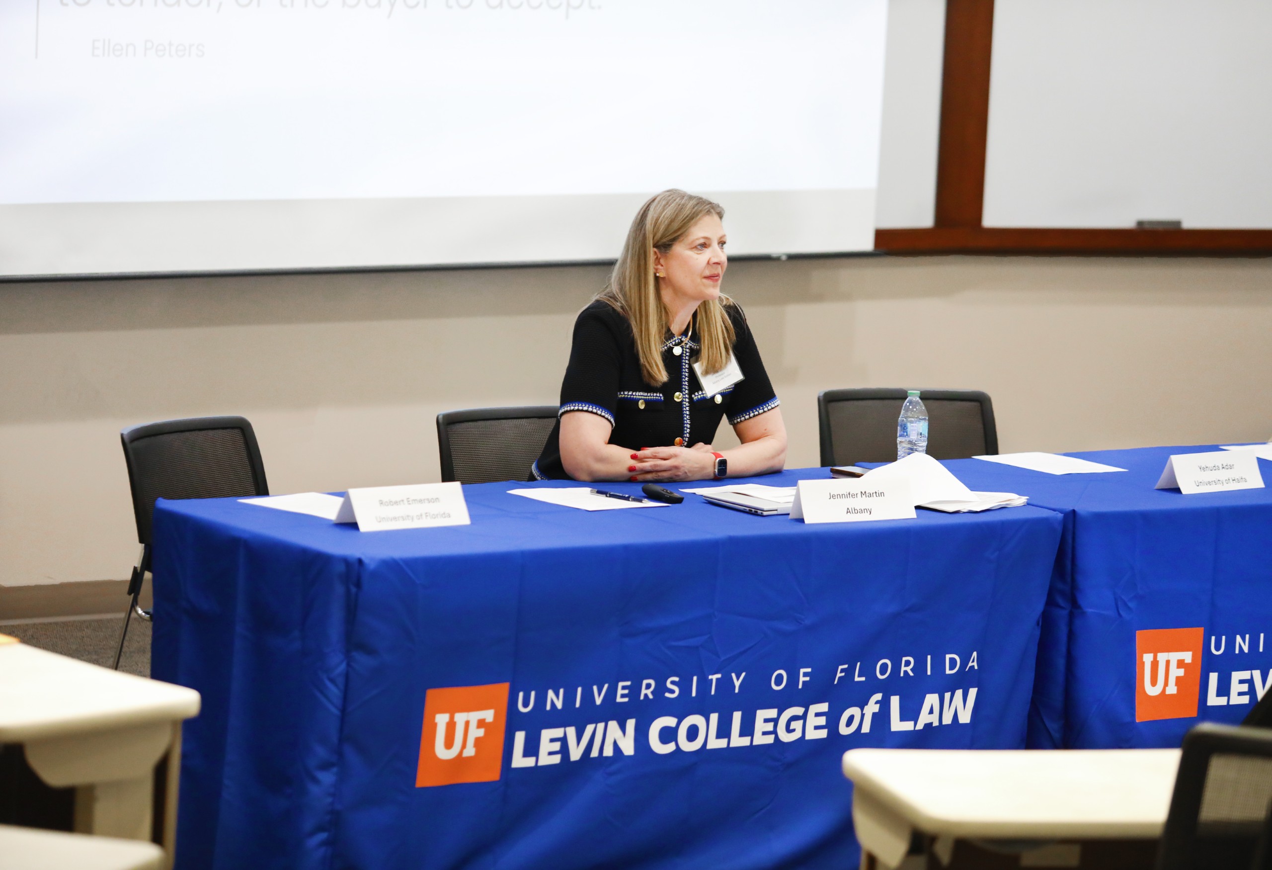 Panelist Jennifer Martin sits at a long table in a lecture room facing the audience. The table is covered with a blue table cloth that has the UF Law logo on it, which says "University of Florida Levin College of Law." There is an empty chair to her left and right.