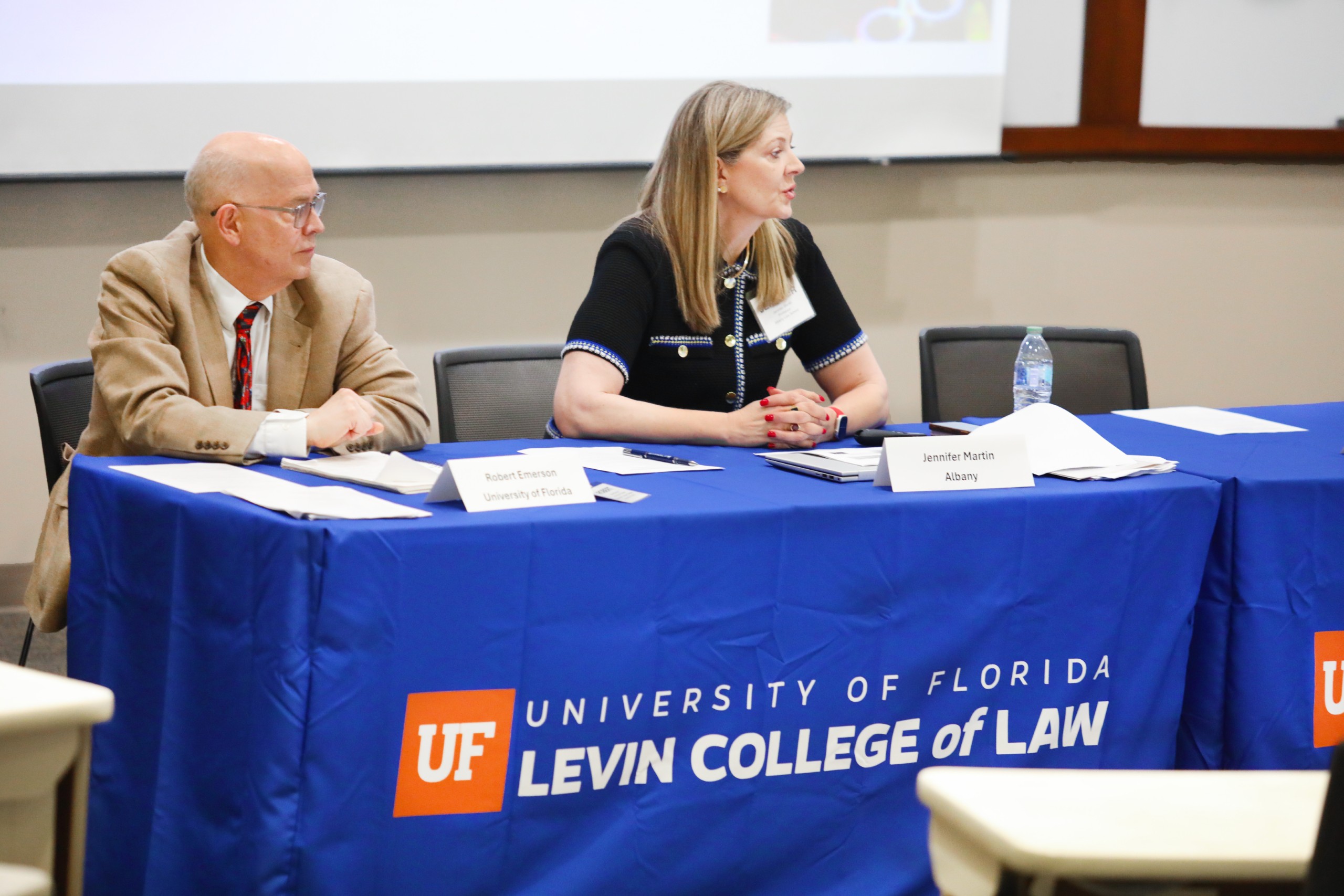 To the left Panelist Robert Emerson (University of Florida) sits at a long table in a lecture room facing the audience. To his right sits Panelist Jennifer Martin, who is speaking to the audience (not pictured) as Emerson listens. The table is covered with a blue table cloth that has the UF Law logo on it, which says "University of Florida Levin College of Law." There is an empty chair to the right of Martin.