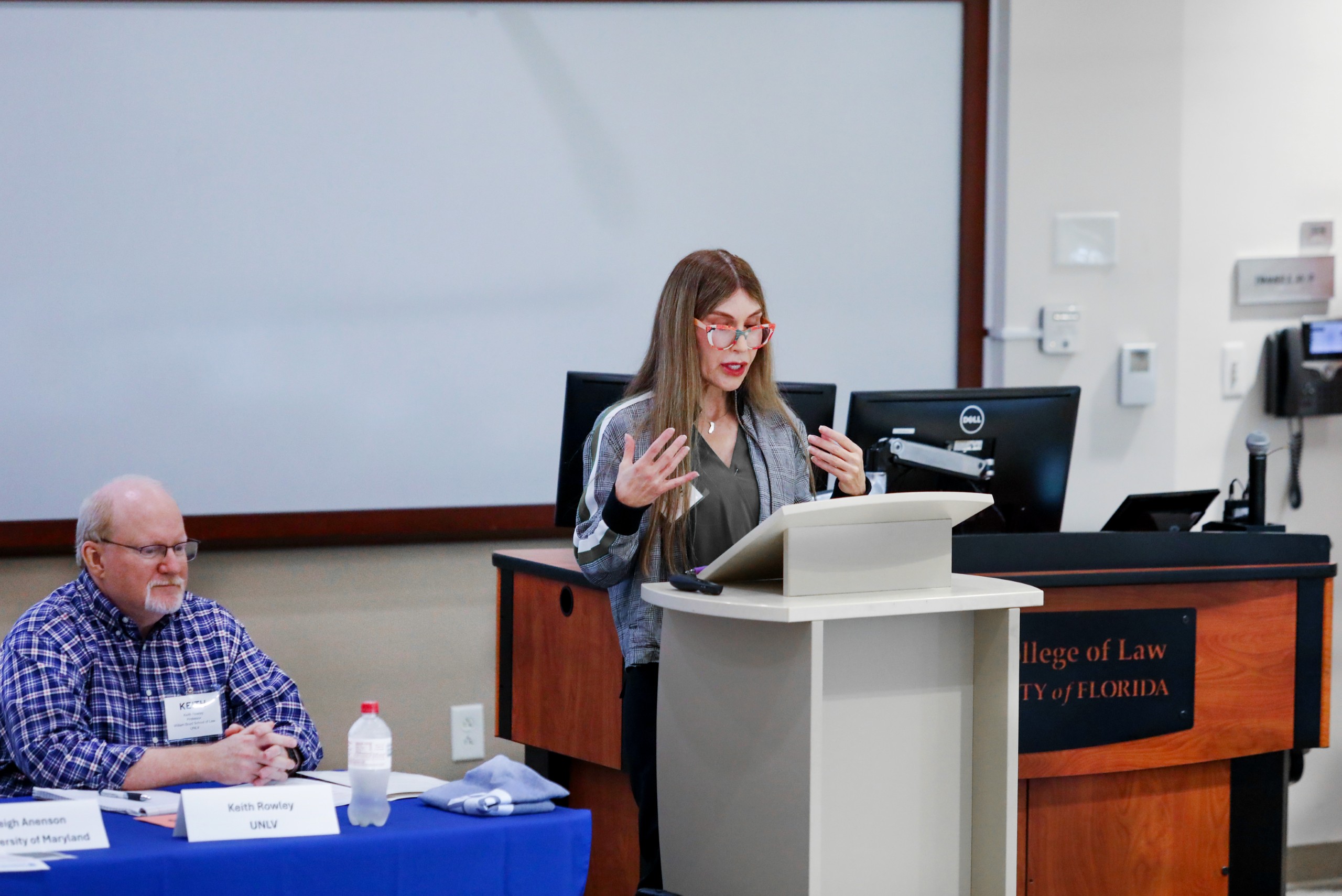 Panel Moderator Keith Rowley (UNLV) sits at the end of a long table in a lecture room facing the audience (not pictured). To his right stands Panelist Leigh Anenson (University of Maryland), who is presenting behind a podium. 