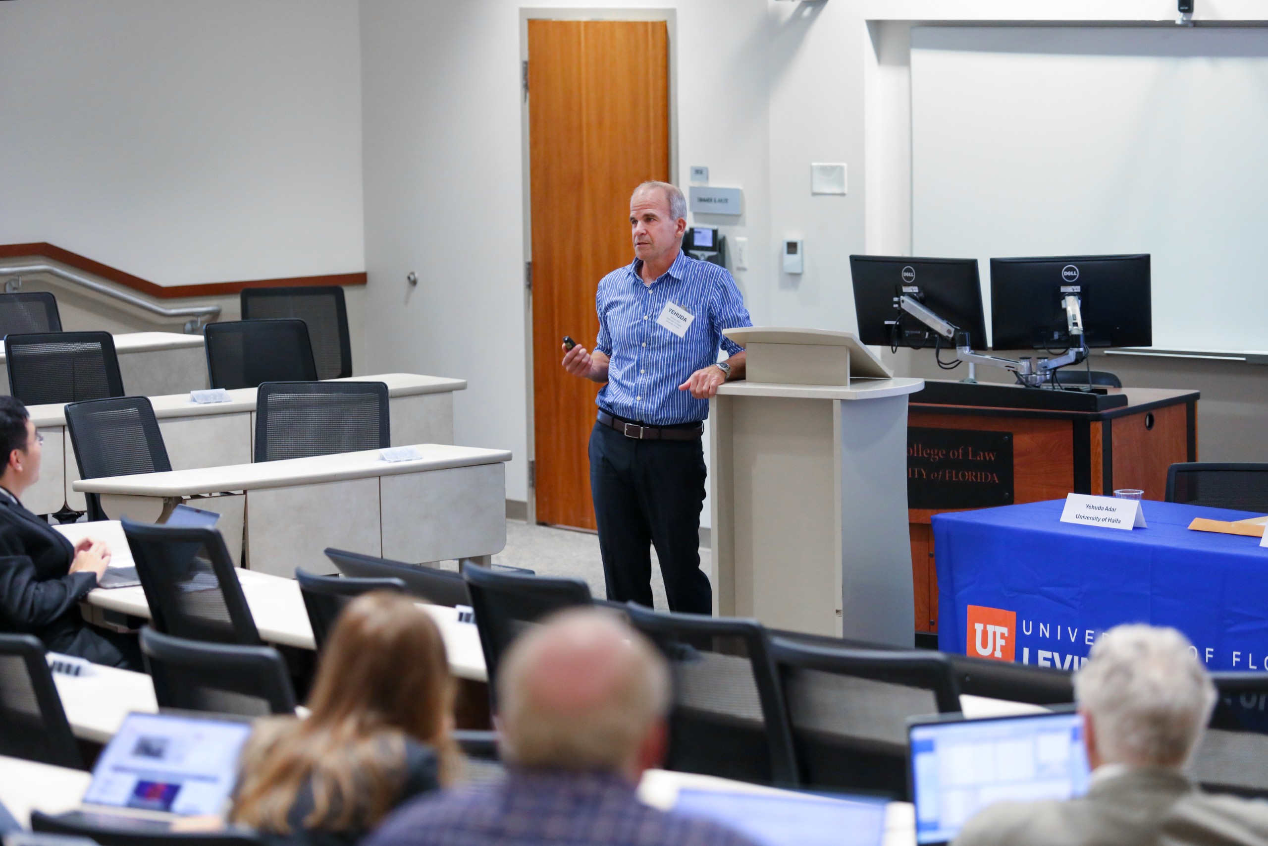 Panelist Yehuda Adar (University of Haifa) stands to the left of long table in a lecture room facing the audience (partially pictured in the foreground). He gestures with his hands while talking to the audience, leaning on a podium to his right.