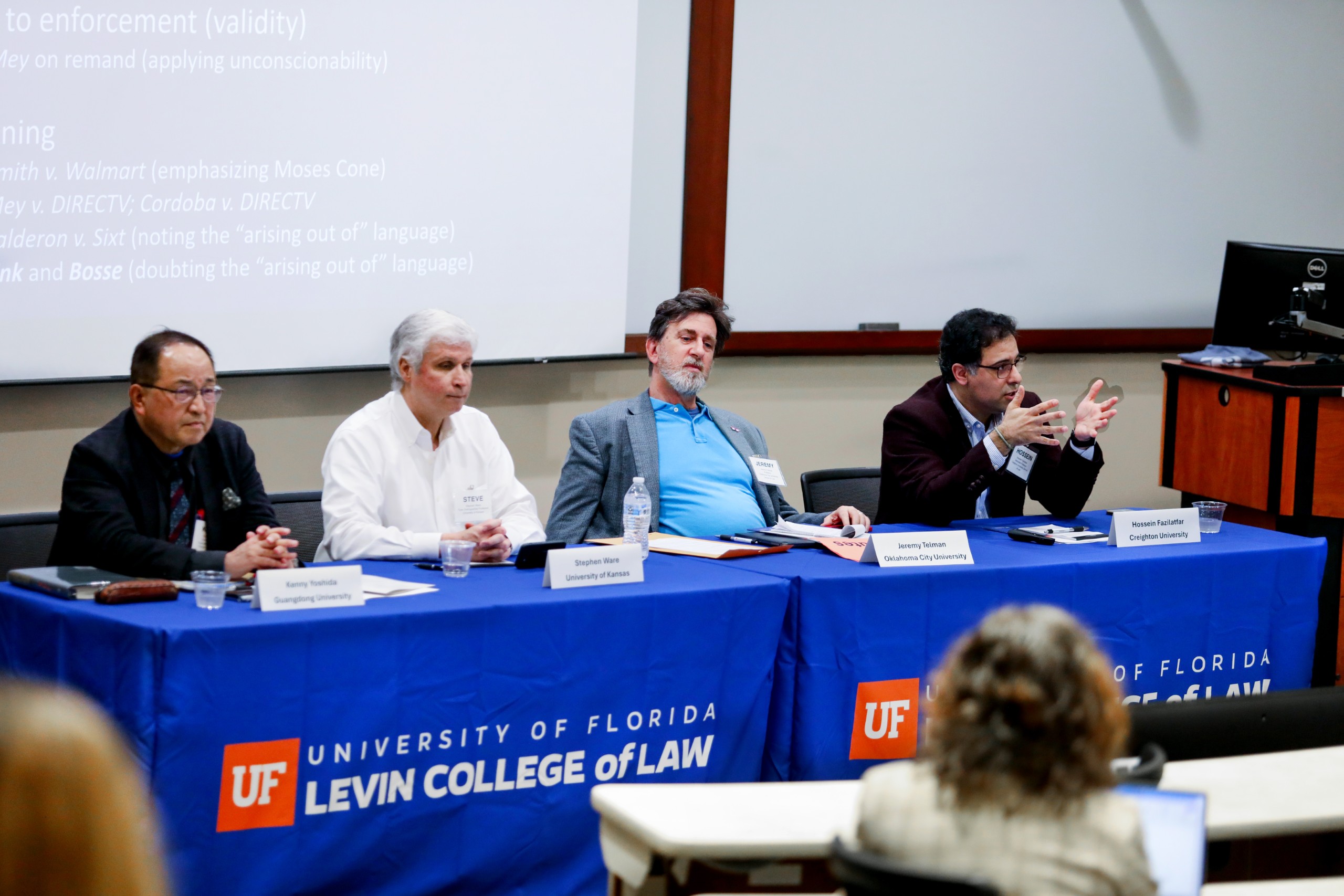 Four people sit at a long table in a lecture room facing the audience who are partially pictured in the foreground. The table is covered with a blue table cloth that has the UF Law logo on it, which says "University of Florida Levin College of Law." From left to right sits Panelist Kenny Yoshida (Guandong University), Panelist Stephen Ware (University of Kansas), Moderator Jeremy Telman (Oklahoma City University), and Panelist Hossein Fazilatfar (Creighton University) who is speaking to the audience while gesturing with his hands.