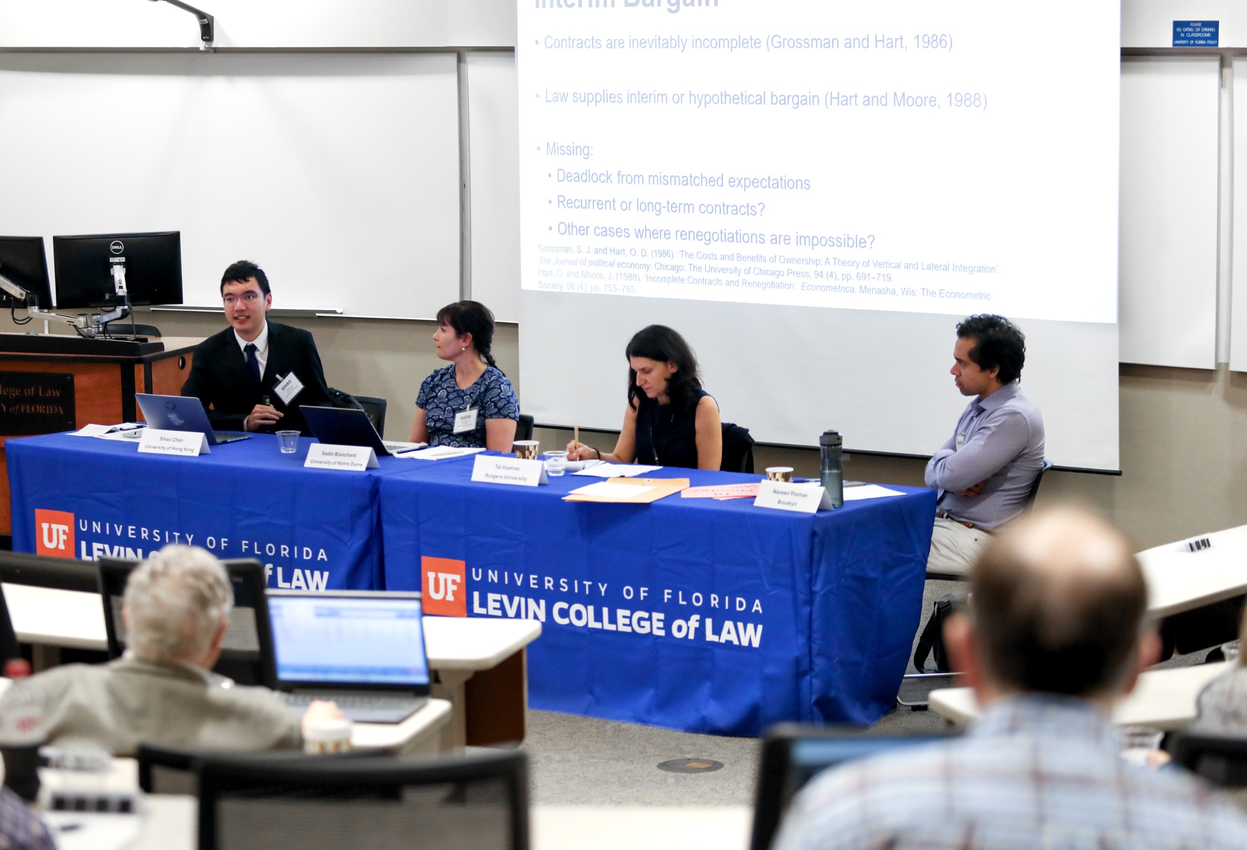 Four people sit at a long table in a lecture room facing the audience who are partially pictured in the foreground. The table is covered with a blue table cloth that has the UF Law logo on it, which says "University of Florida Levin College of Law." From left to right sits Panelist Sihao Chen (University of Hong Kong), Sadie Blanchard (University of Notre Dame), Tal Kastner (Rutgers), and Moderator Naveen Thomas (Brooklyn). Behind them is a slide projection with bullet points.