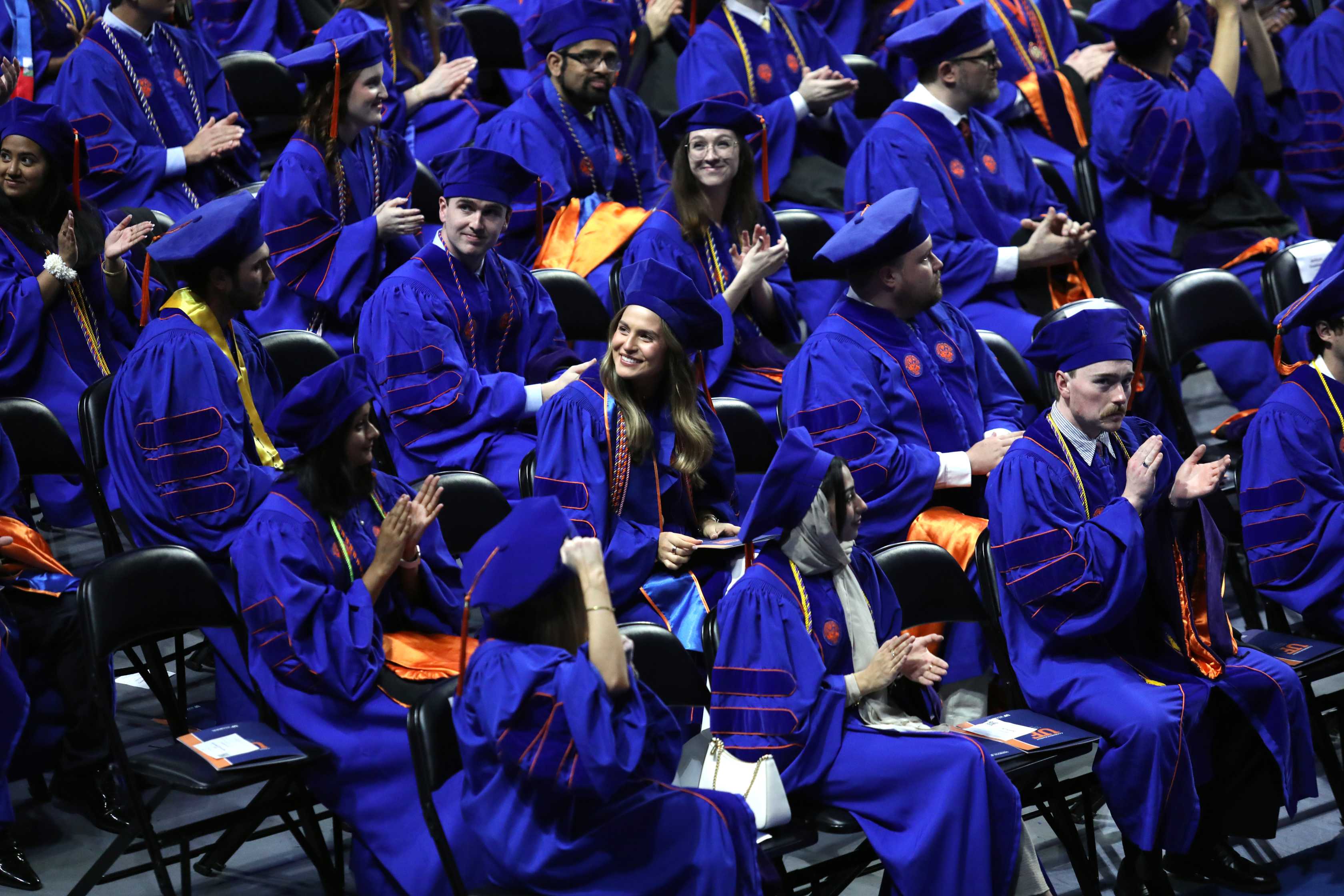 A group of graduates in caps and gowns sits together, some clapping and smiling during a ceremony. The audience is arranged in rows, with a mix of expressions reflecting pride and celebration.
