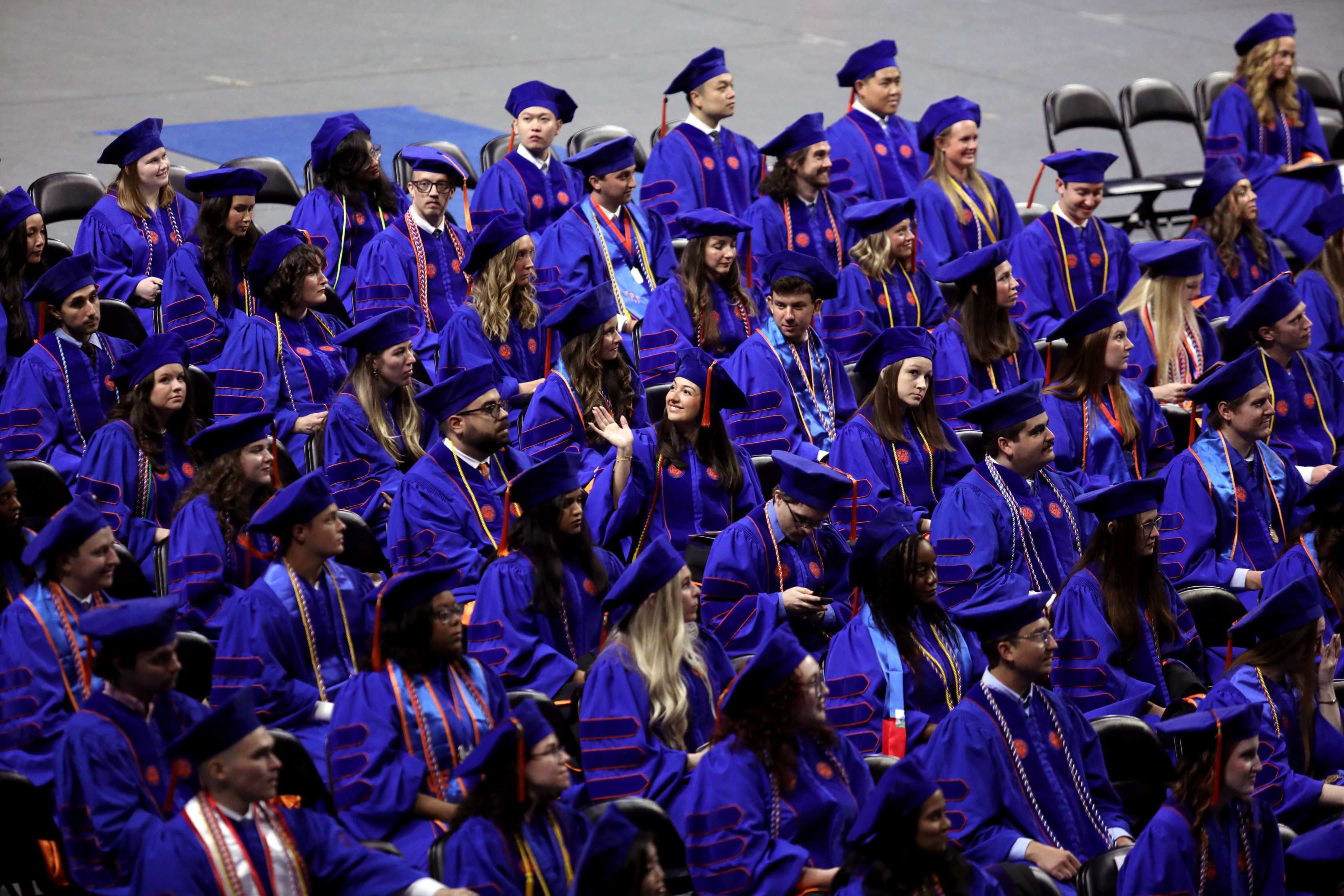 A gathering of graduates in blue caps and gowns is seated in a large hall. The audience is focused and engaged in the ceremony, with some individuals smiling and raising hands.
