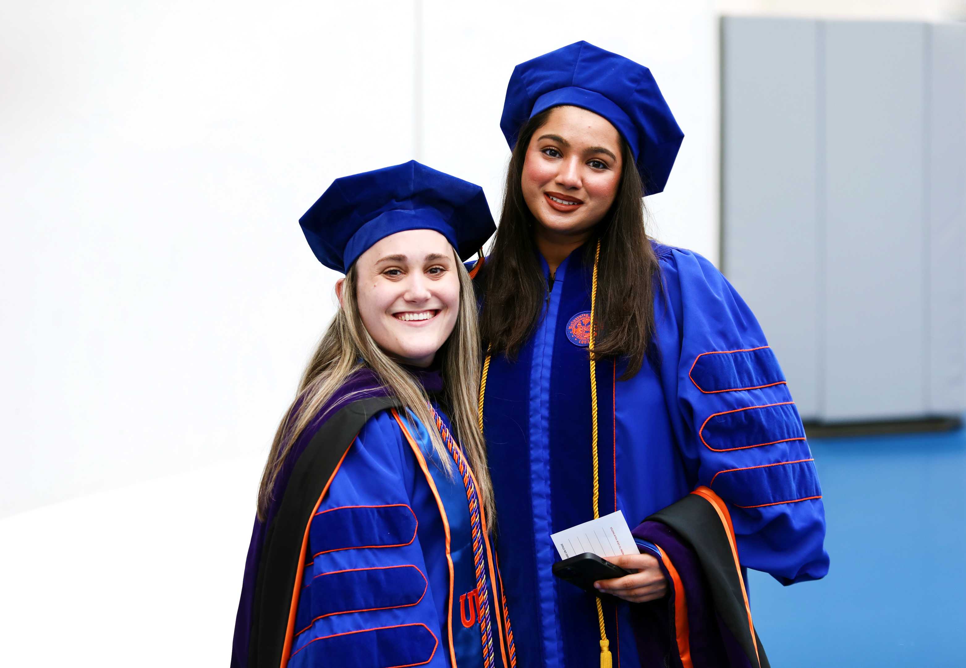 Two individuals in academic regalia pose together, smiling. They are wearing blue gowns and caps. The background is a plain, light-colored wall.

