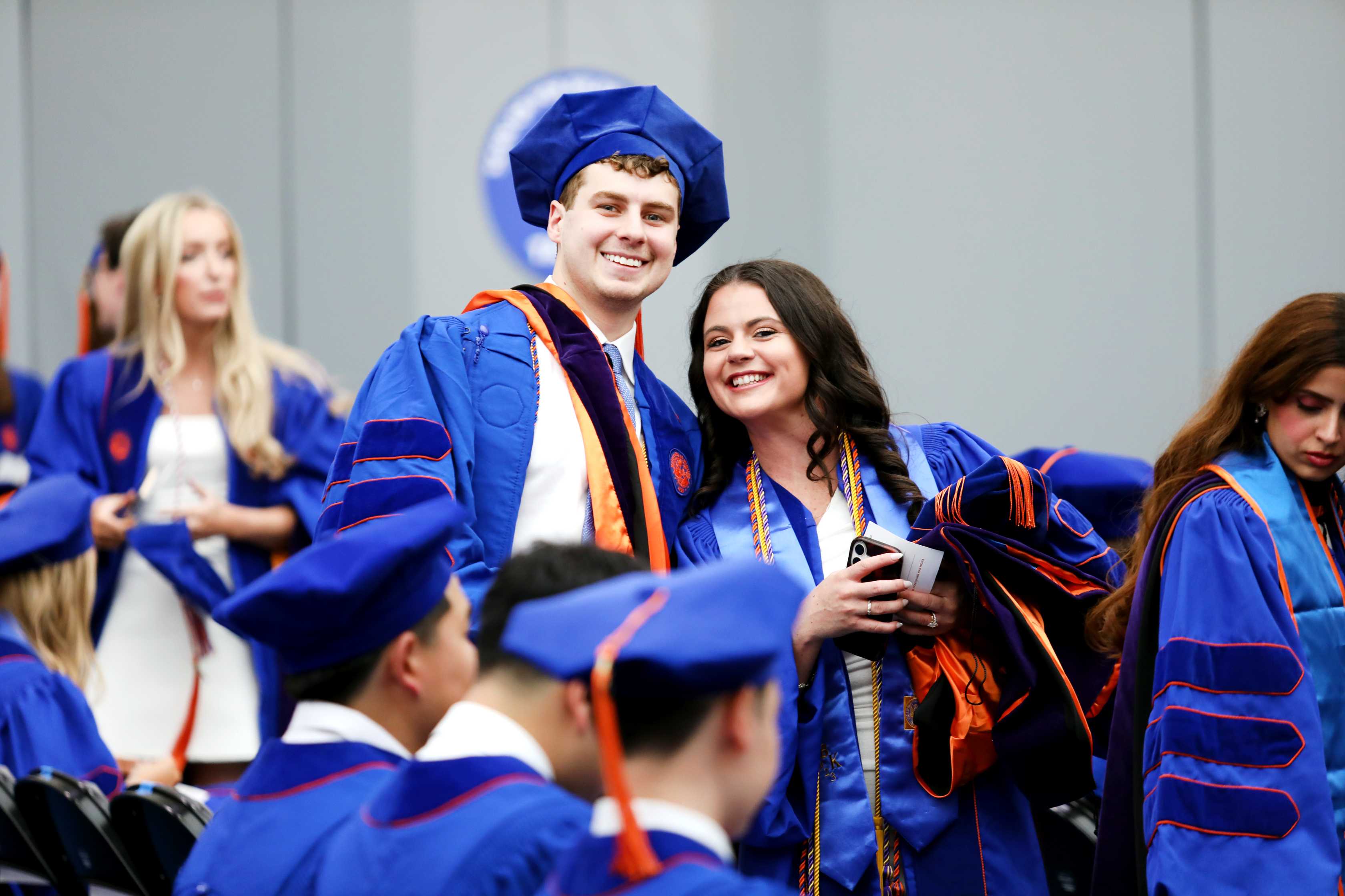 A group of graduates in blue robes and caps smiles and poses for a photo in a ceremony setting, with many others in the background seated.