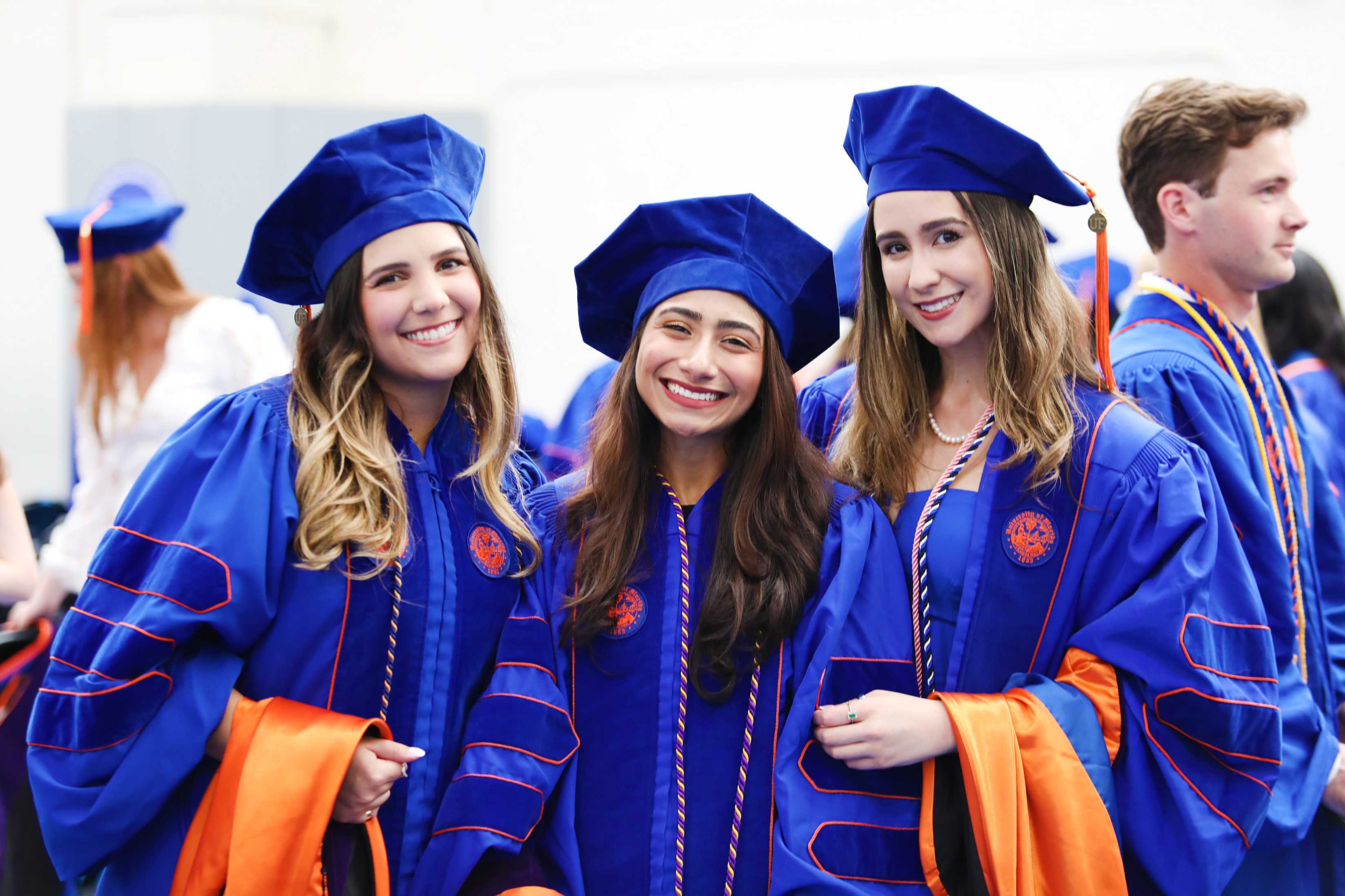 Three individuals in blue graduation gowns and caps smile together. They are surrounded by others in similar attire, celebrating a graduation event.
