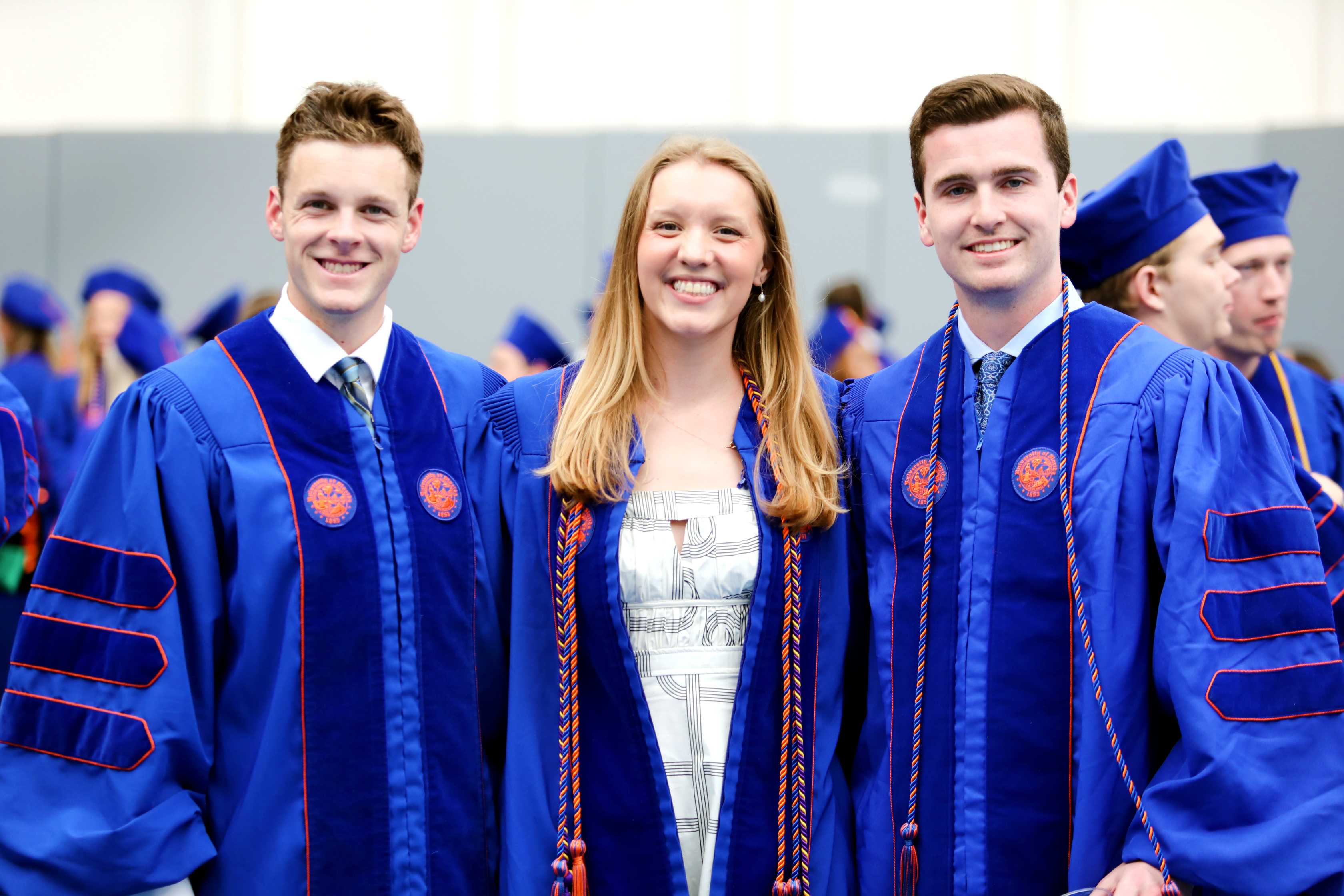 A group of graduates in blue robes and caps smiles and poses for a photo in a ceremony setting, with many others in the background seated.