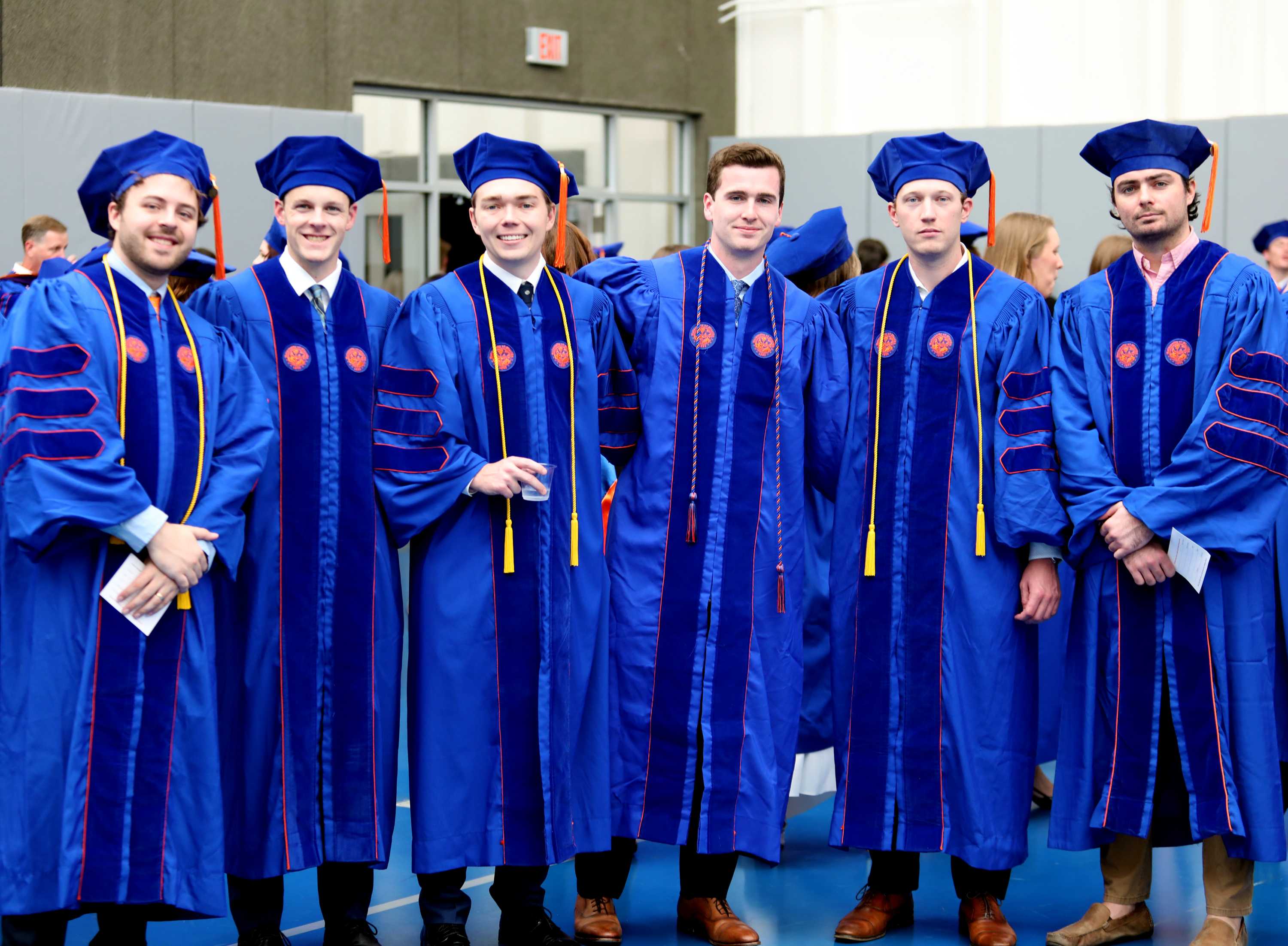 Group of six individuals posing together, all wearing blue graduation gowns with colorful accents. They stand in a celebratory manner, smiling, in an indoor setting.
