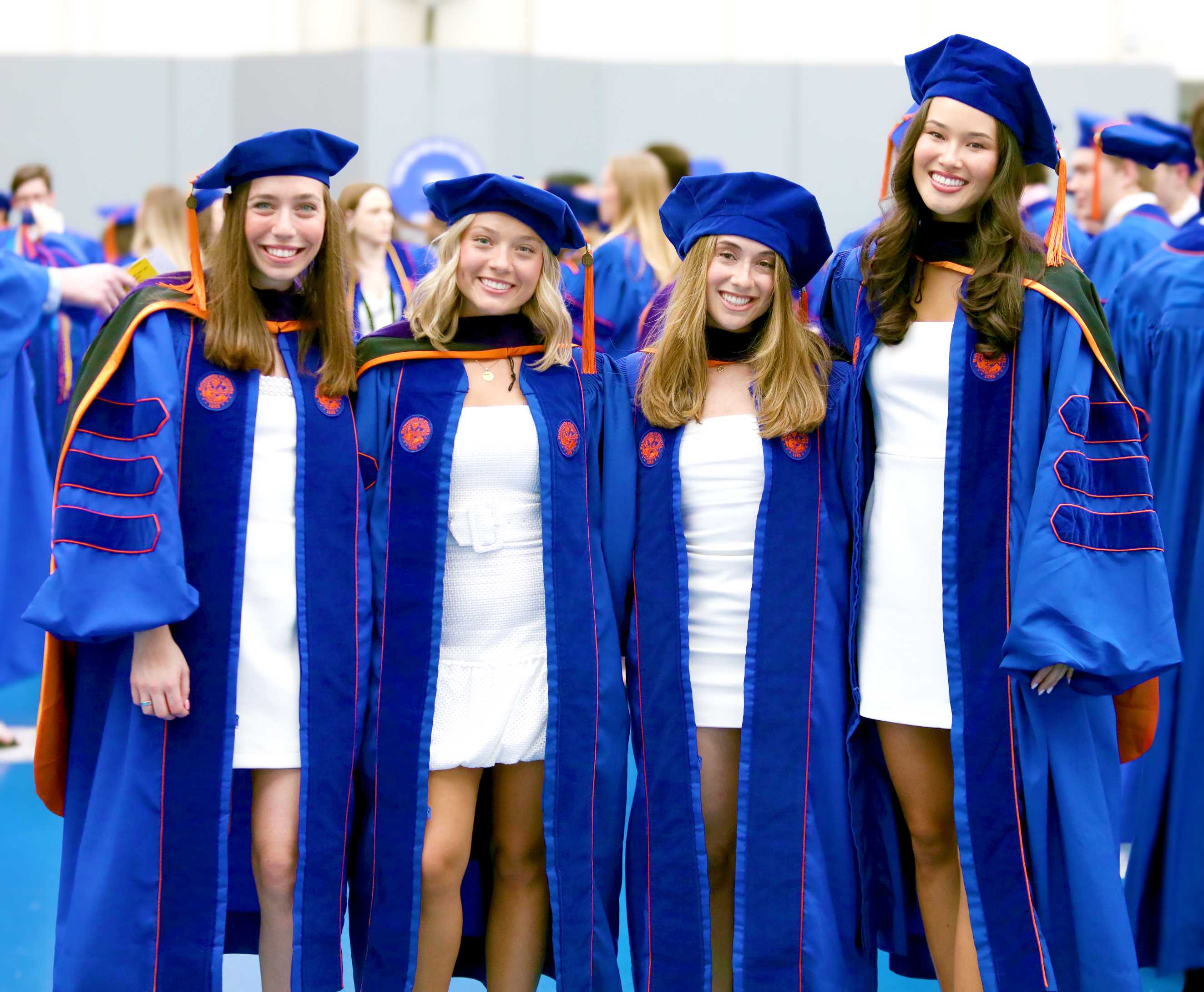 Four individuals stand together, smiling, in blue graduation gowns and caps. They are in a celebratory environment, with others visible in the background.
