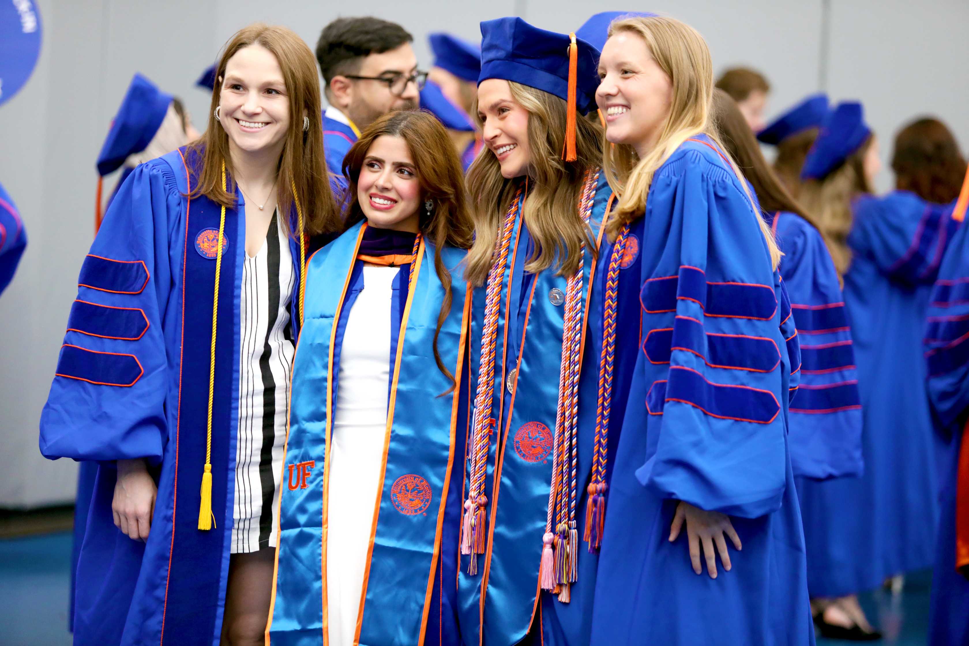Four individuals in blue graduation gowns and caps are standing together, smiling for a photo. The background features other graduates in similar attire, creating a celebratory atmosphere.
