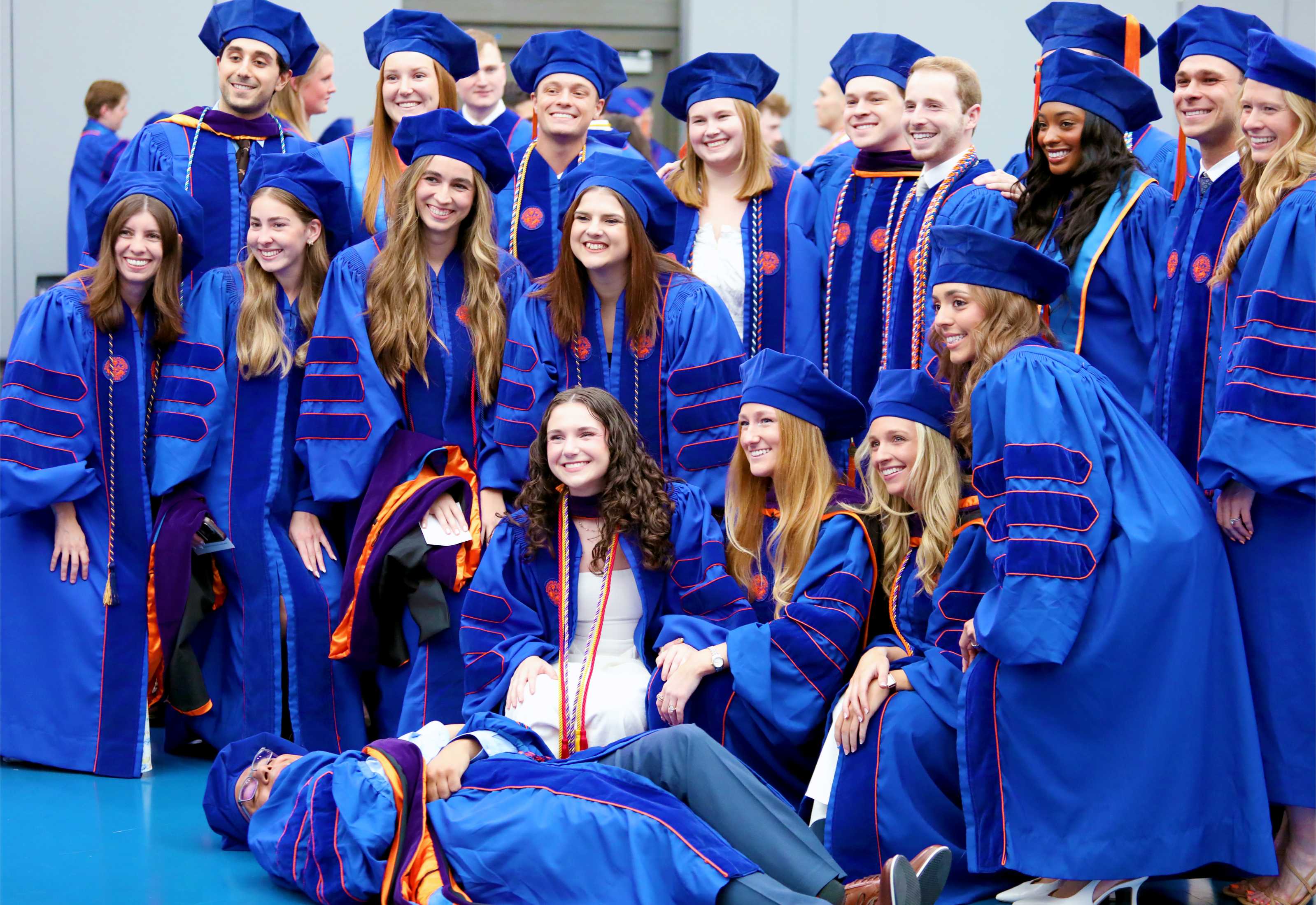 A group of graduates in blue academic gowns and caps poses together, smiling and celebrating their achievement. One individual is lying on the ground in front of the group, adding a playful element to the photo.