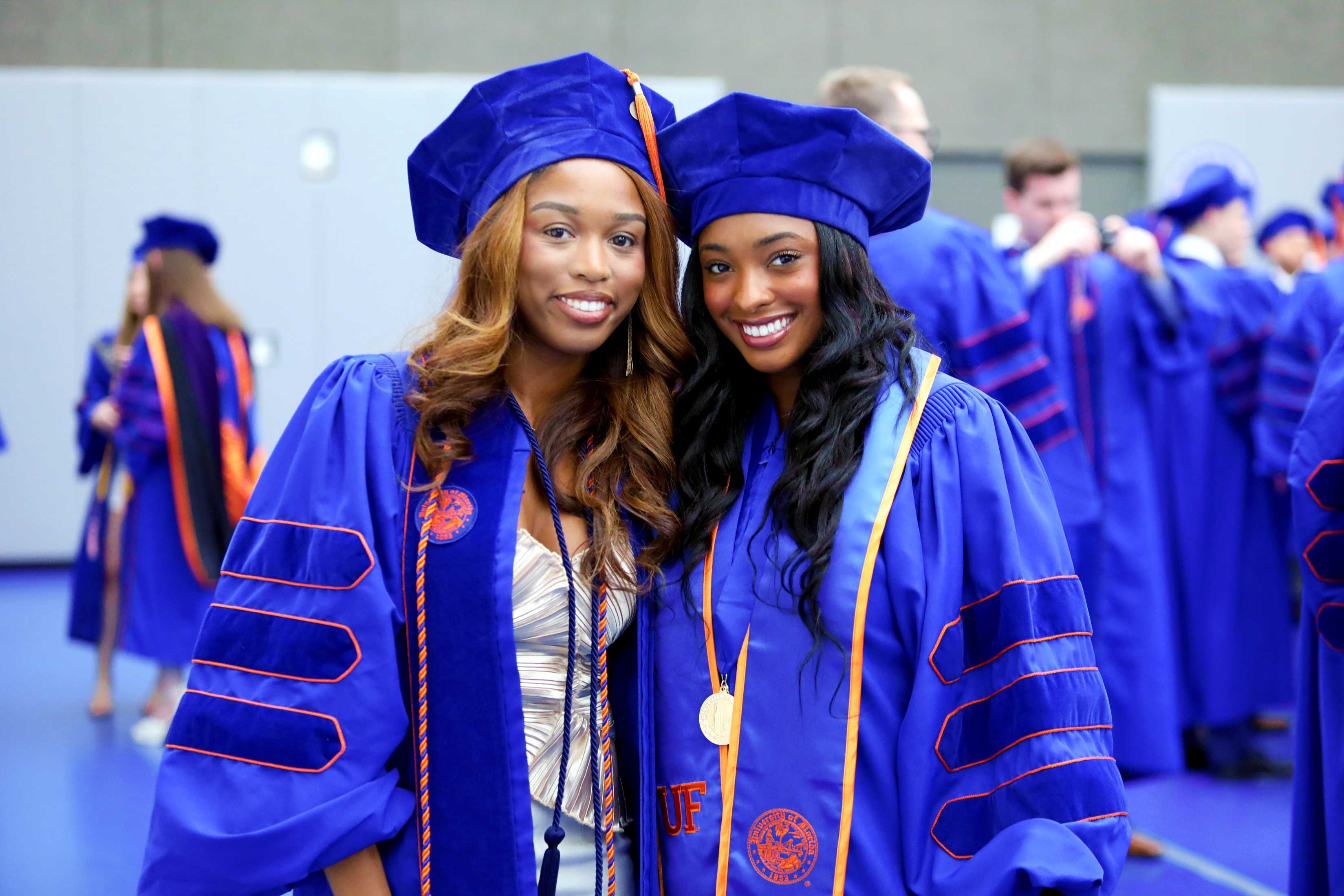 Two individuals wearing blue graduation gowns and caps are smiling and posing together in a large assembly area filled with other graduates in the background.
