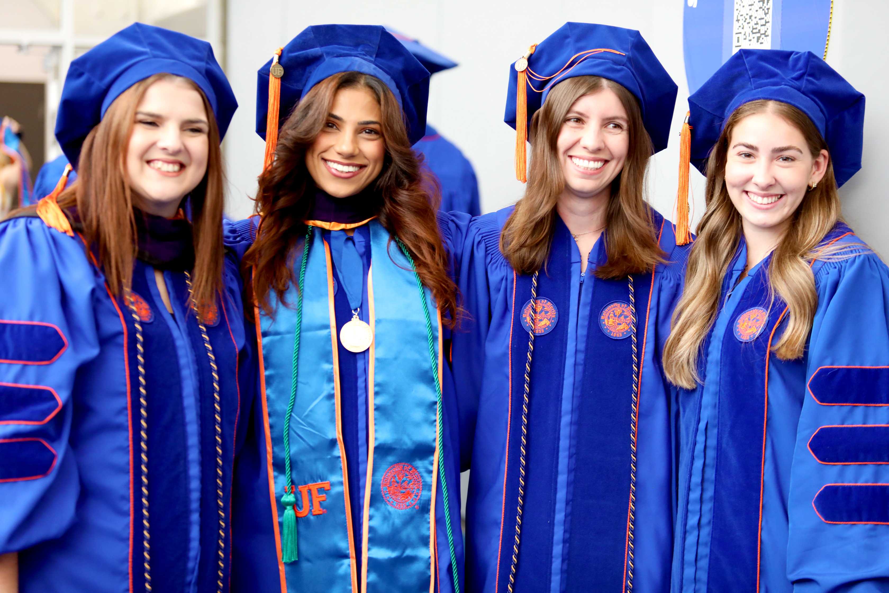 Four individuals are smiling together, dressed in blue graduation robes and caps. They are positioned closely, celebrating a graduation event.
