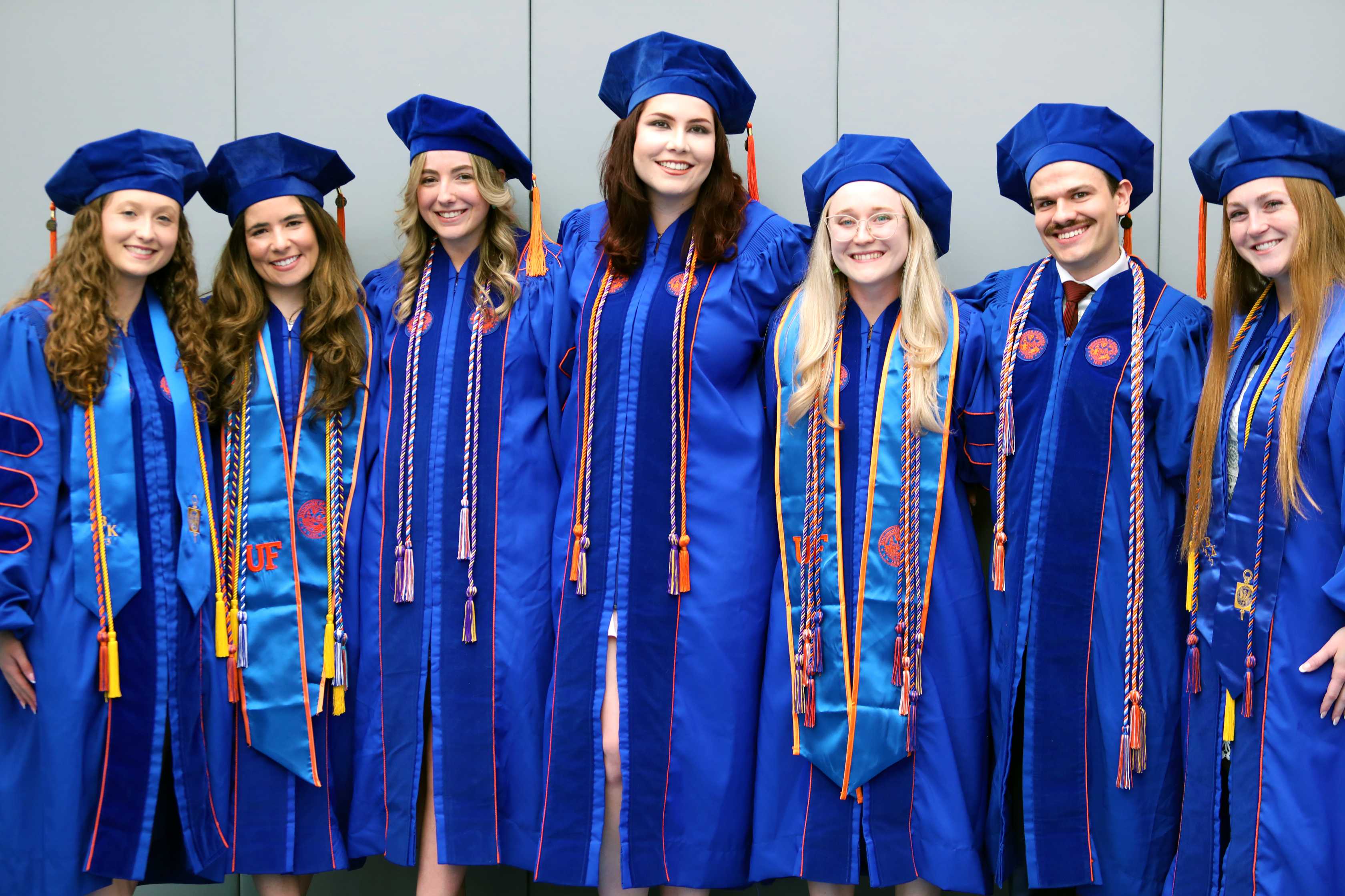 Seven individuals in blue graduation gowns and caps stand together, smiling. They are positioned closely, showcasing their attire and graduation cords. The background is neutral and unobtrusive.
