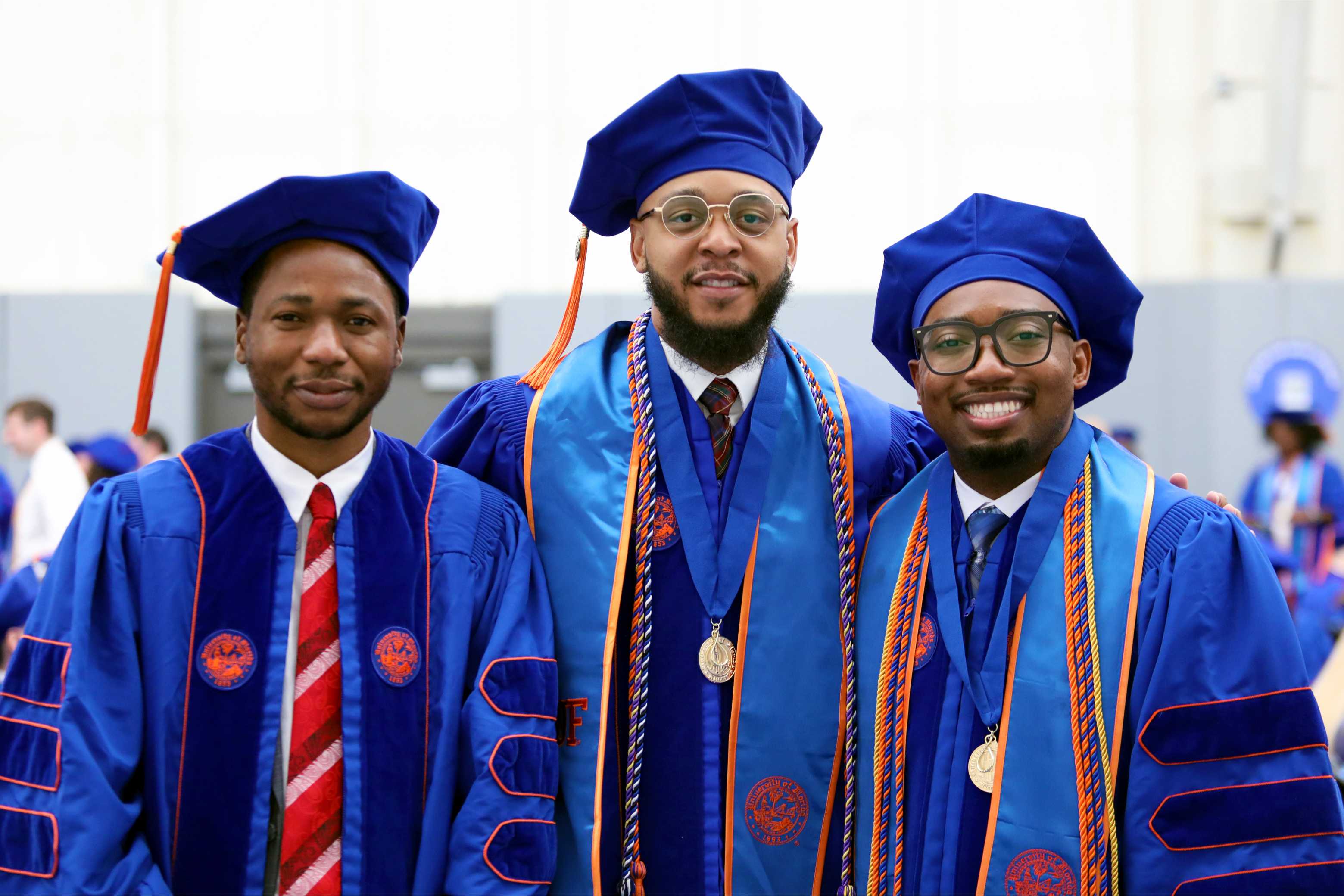 Three graduates stand together, all wearing blue academic regalia and caps. One person is in the center, smiling broadly, while the others are on either side, also smiling. The background features blurred figures in similar attire, suggesting a graduation ceremony setting.