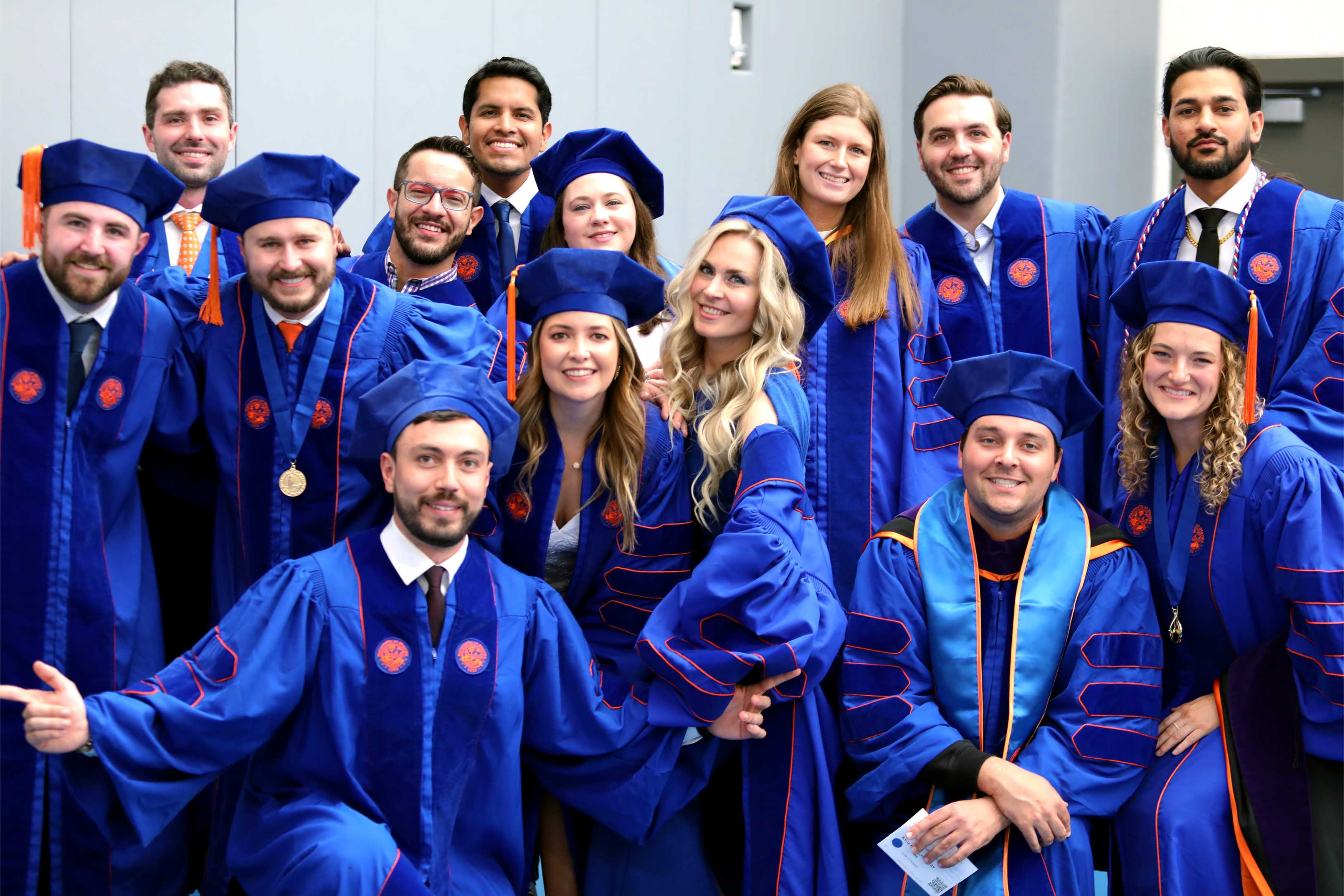A group of graduates in blue gowns and caps poses together, smiling and celebrating their achievement. The setting suggests a graduation ceremony, with individuals standing and kneeling in two rows.
