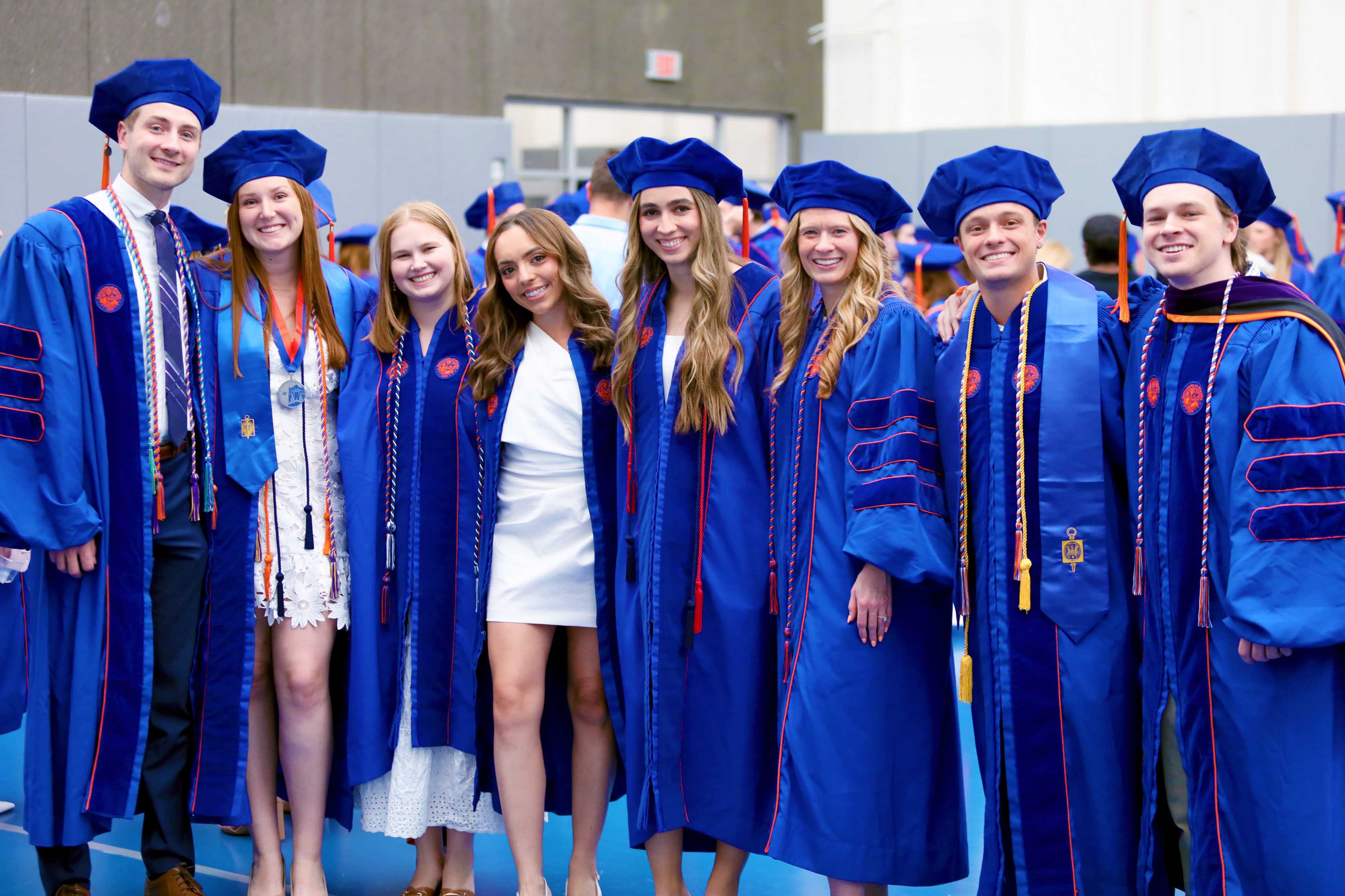 A group of eight graduates in blue academic regalia stands together, smiling in a celebratory pose. They are inside a building with others in the background, wearing similar attire.
