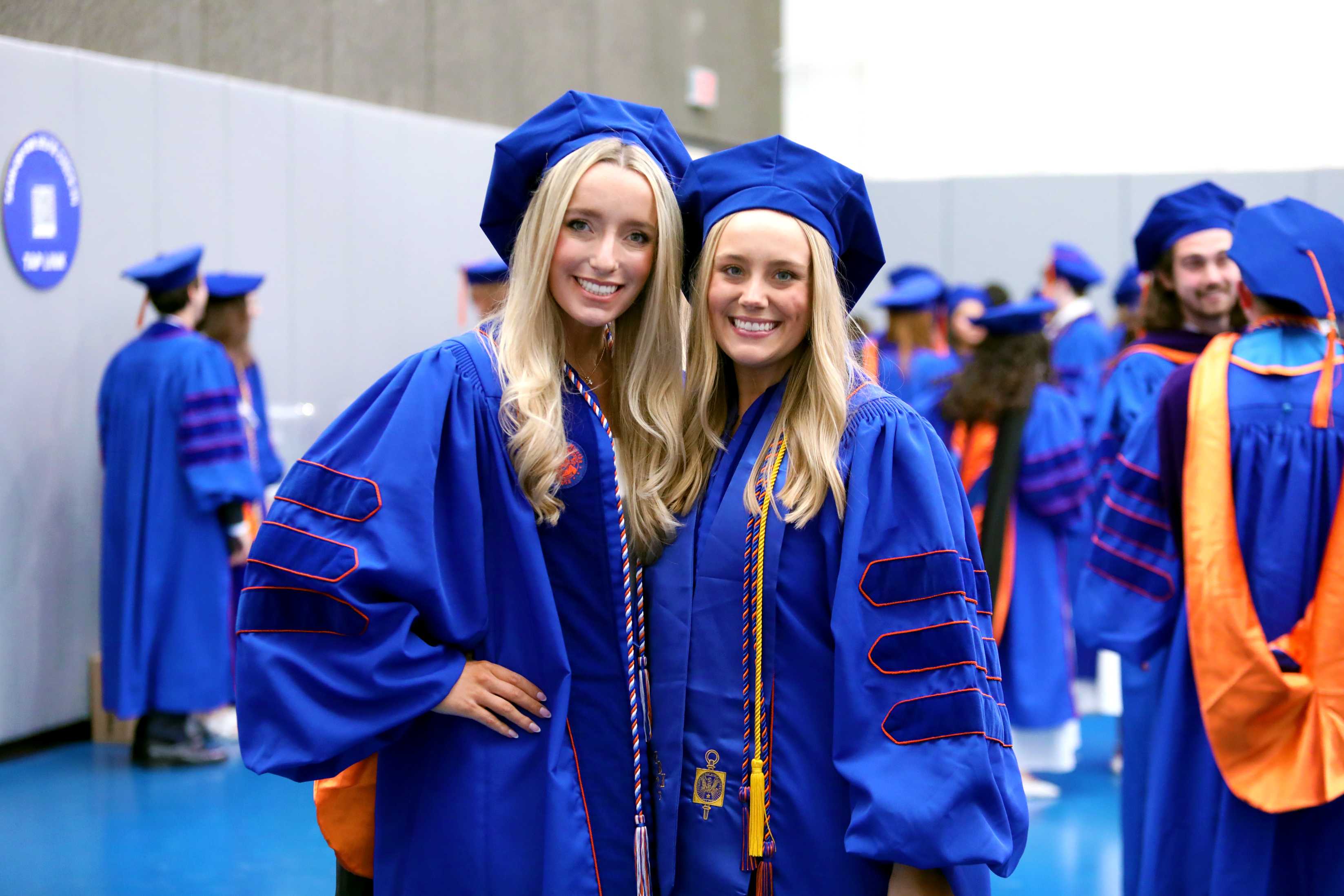 Two individuals smile and pose together in blue graduation robes with caps. They are in a crowded setting with others dressed similarly in the background.