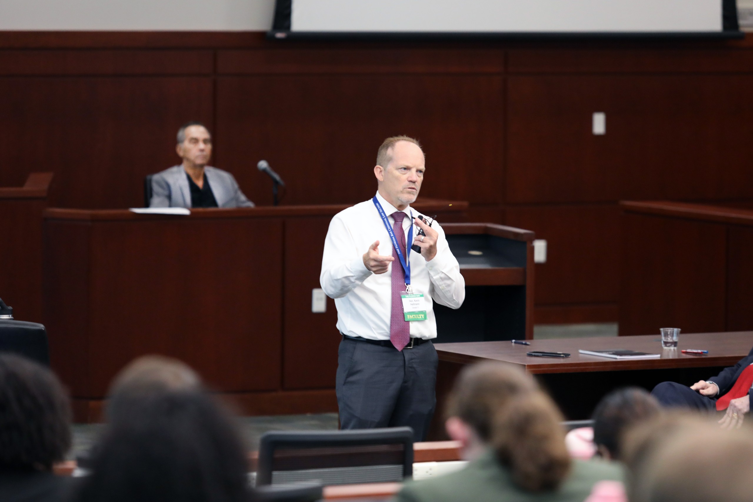 A person stands behind a lectern, speaking to an audience. Another individual is seated in the background. The room has a dark wood paneling and a screen is visible on the wall.