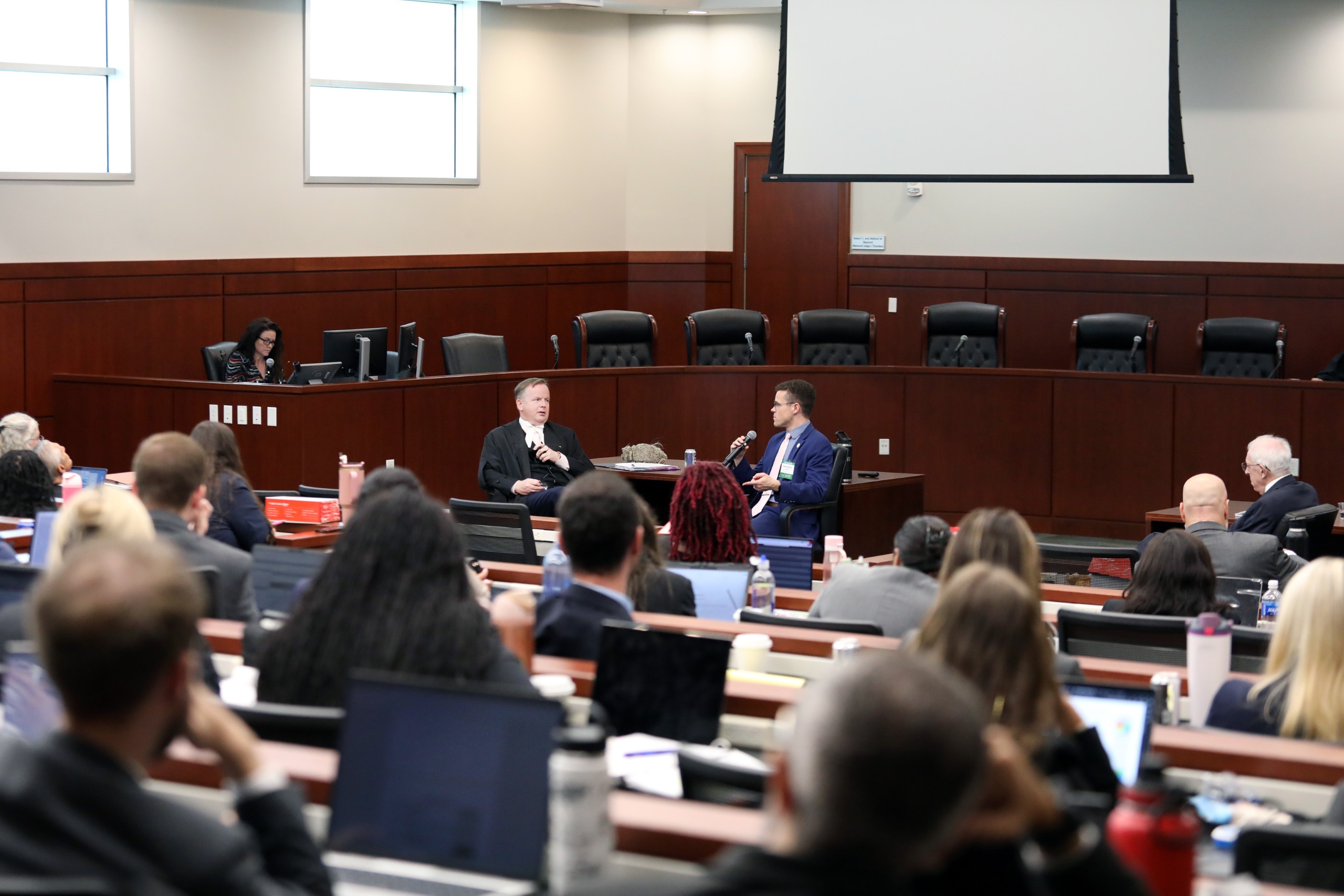 A speaker sits behind a lectern, engaged in conversation with another individual in a large room filled with an audience. A person is visible at a computer in the background. The setting appears to be a formal event or discussion.
