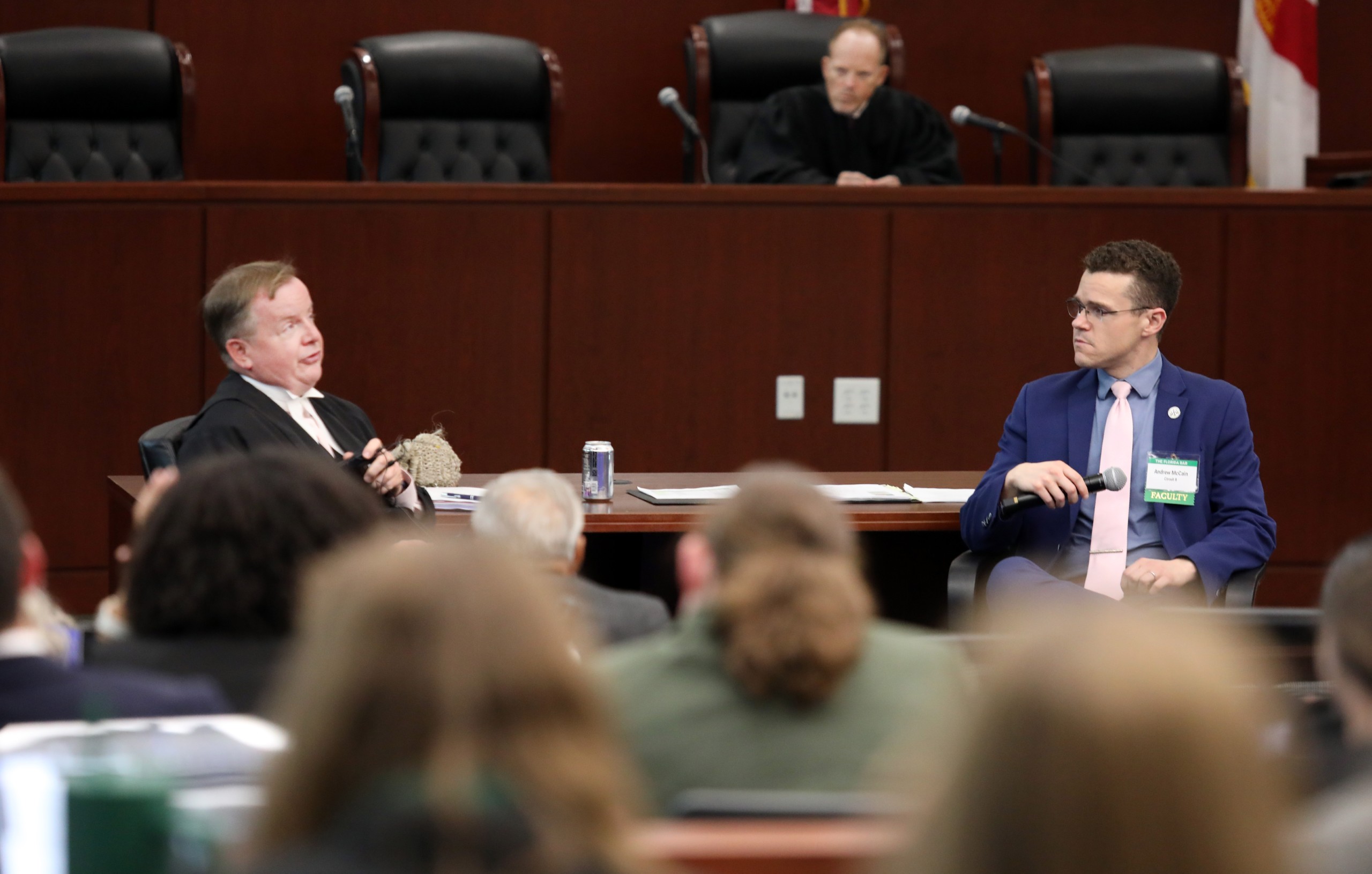 A speaker is seated behind a lectern, engaging in discussion with another individual at a table. In the background, a judge is present on an elevated bench, with an audience visible in the foreground.