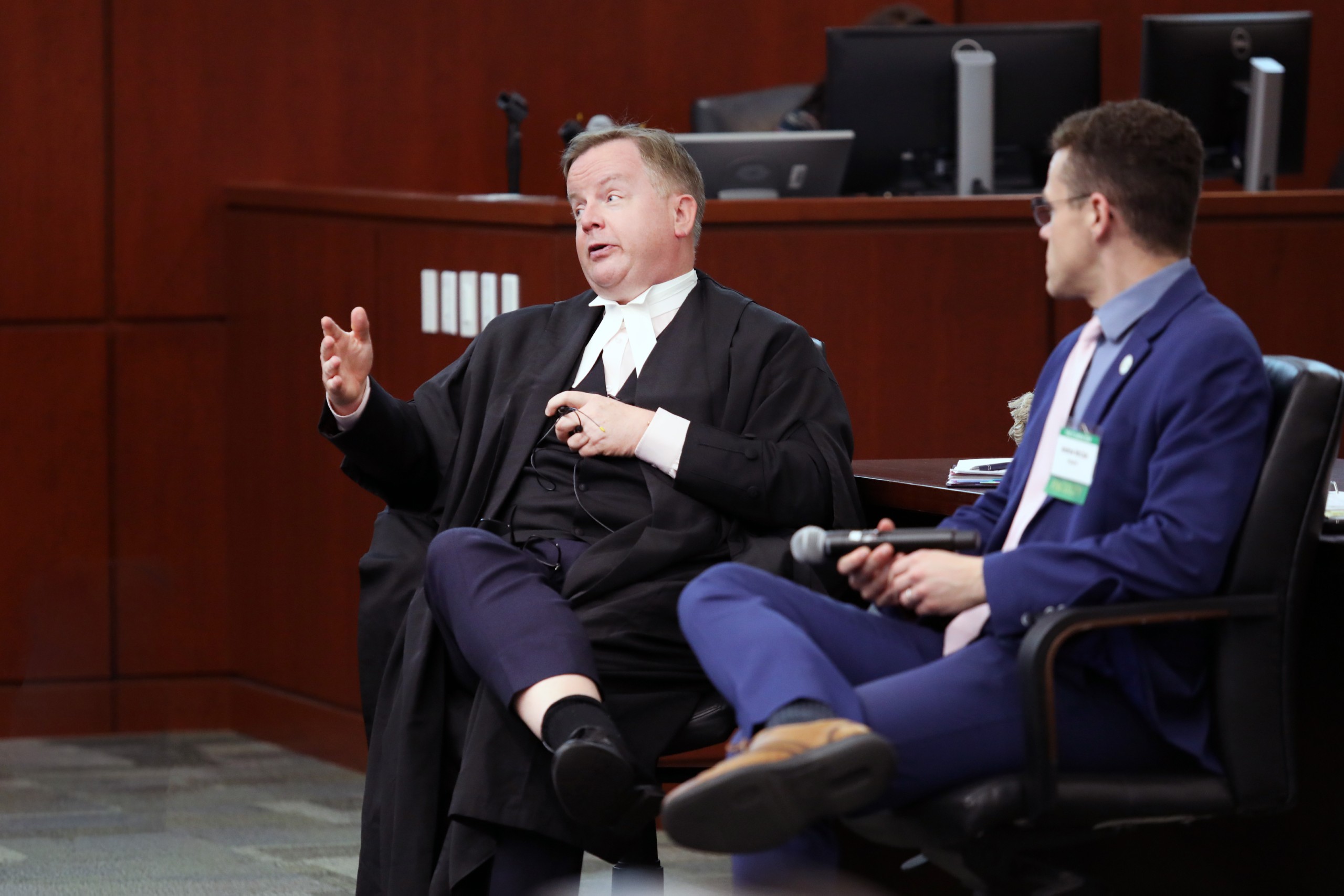 Two individuals are seated in a conference setting. One person is speaking, gesturing with a hand, while the other listens attentively. Both are seated in chairs, and there is a lectern in the background. The room features wooden accents and digital equipment.
