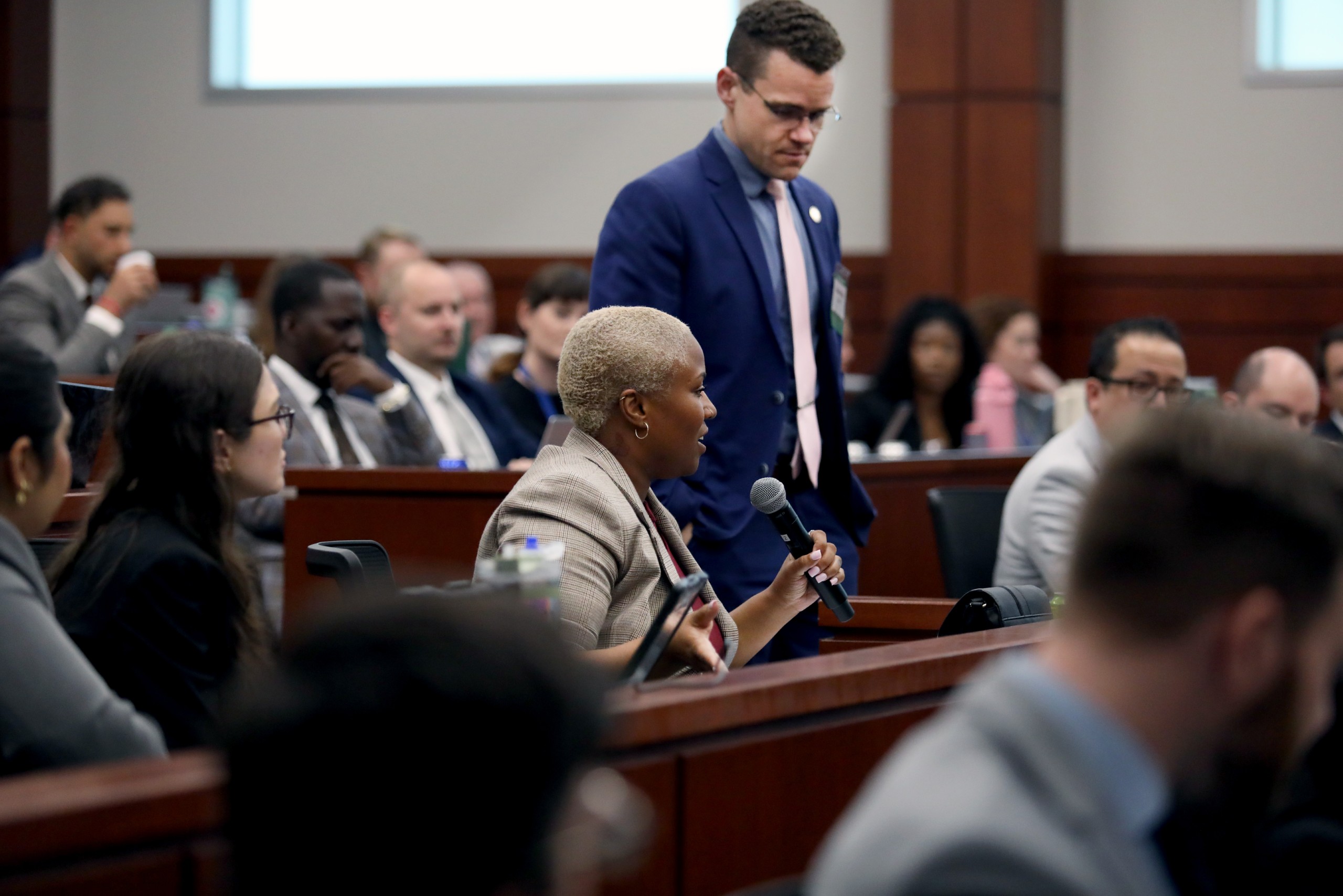 A person stands in a meeting room holding a microphone while speaking, with an audience seated around them, some looking attentively. Another individual in a suit stands nearby, observing the discussion.