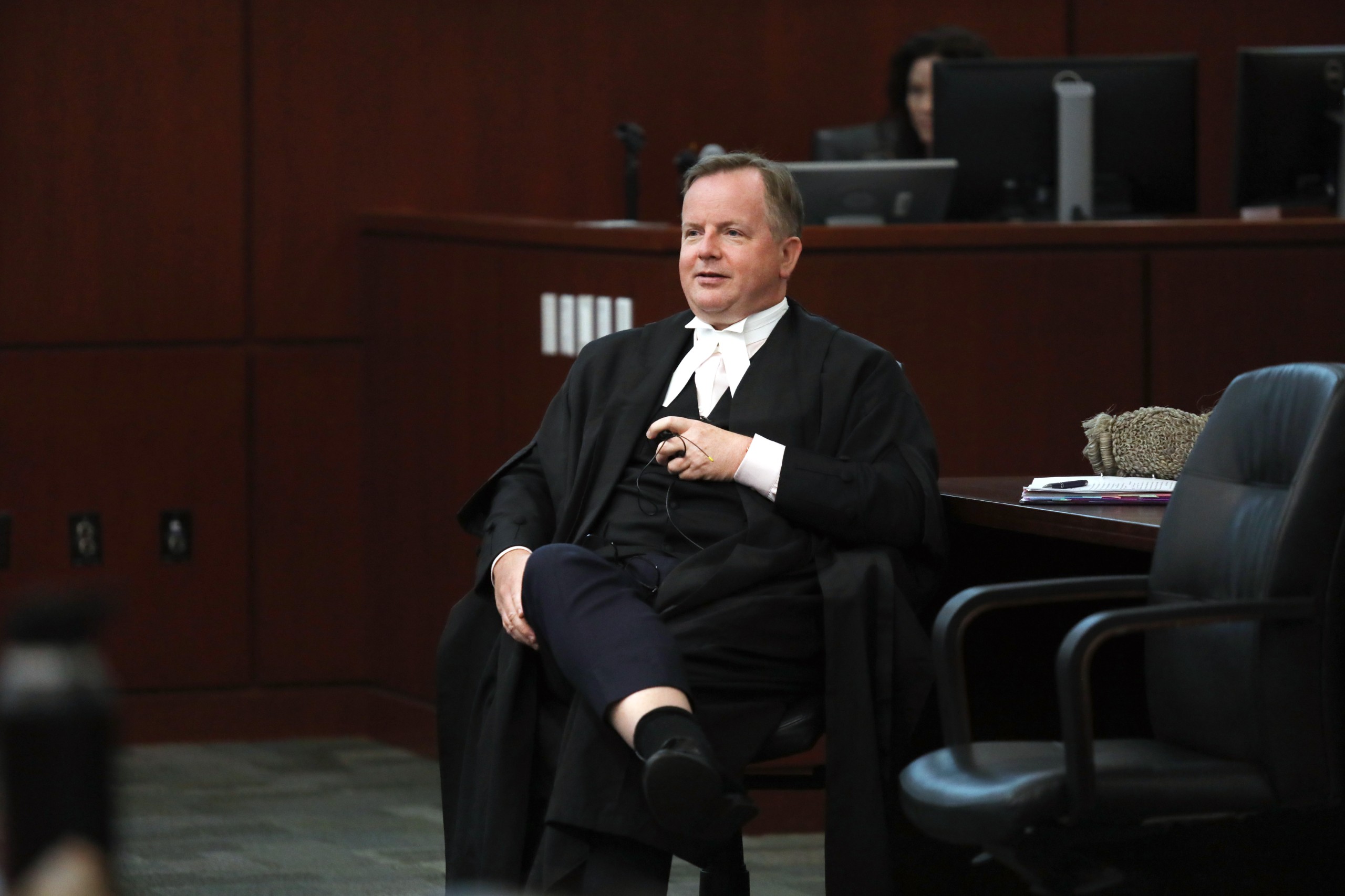 An individual seated in a courtroom, dressed in legal attire, appears relaxed while holding an object in their hand. The background includes desks and equipment typically found in a legal setting.