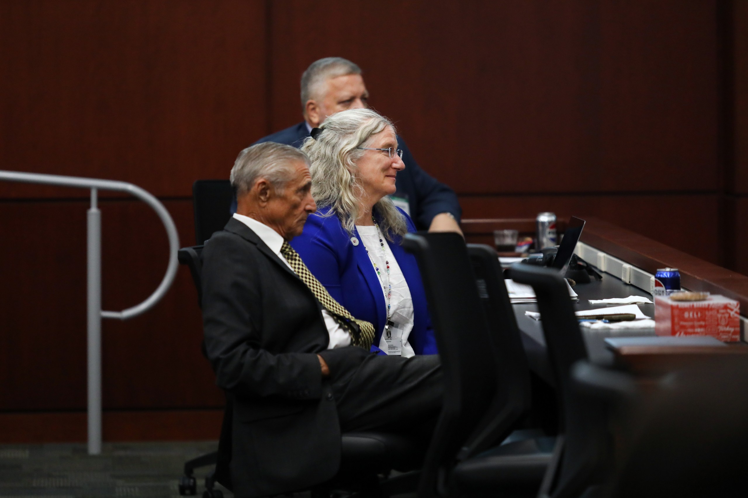 A group of individuals is seated in a courtroom, with one person leaning back in a chair and another smiling while listening. A table with various items, including drinks and snacks, is visible in the foreground.