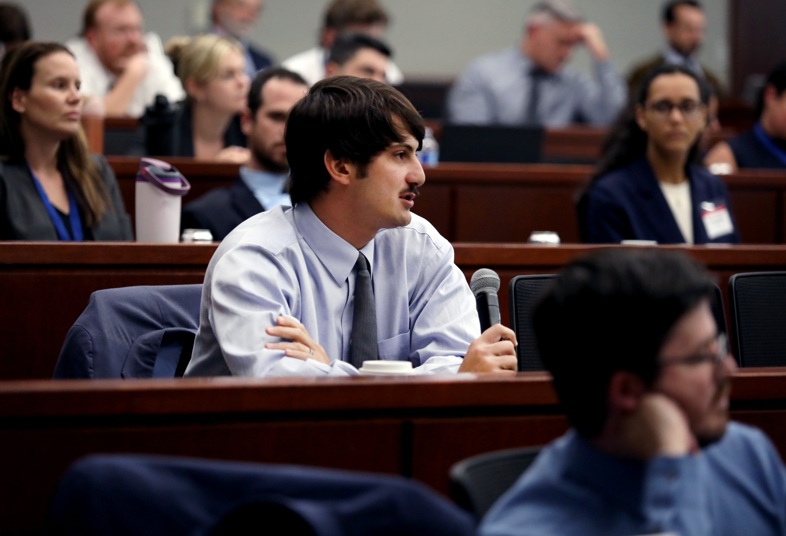 A person sitting in a meeting room raises a hand while holding a microphone. In the background, other attendees listen attentively.