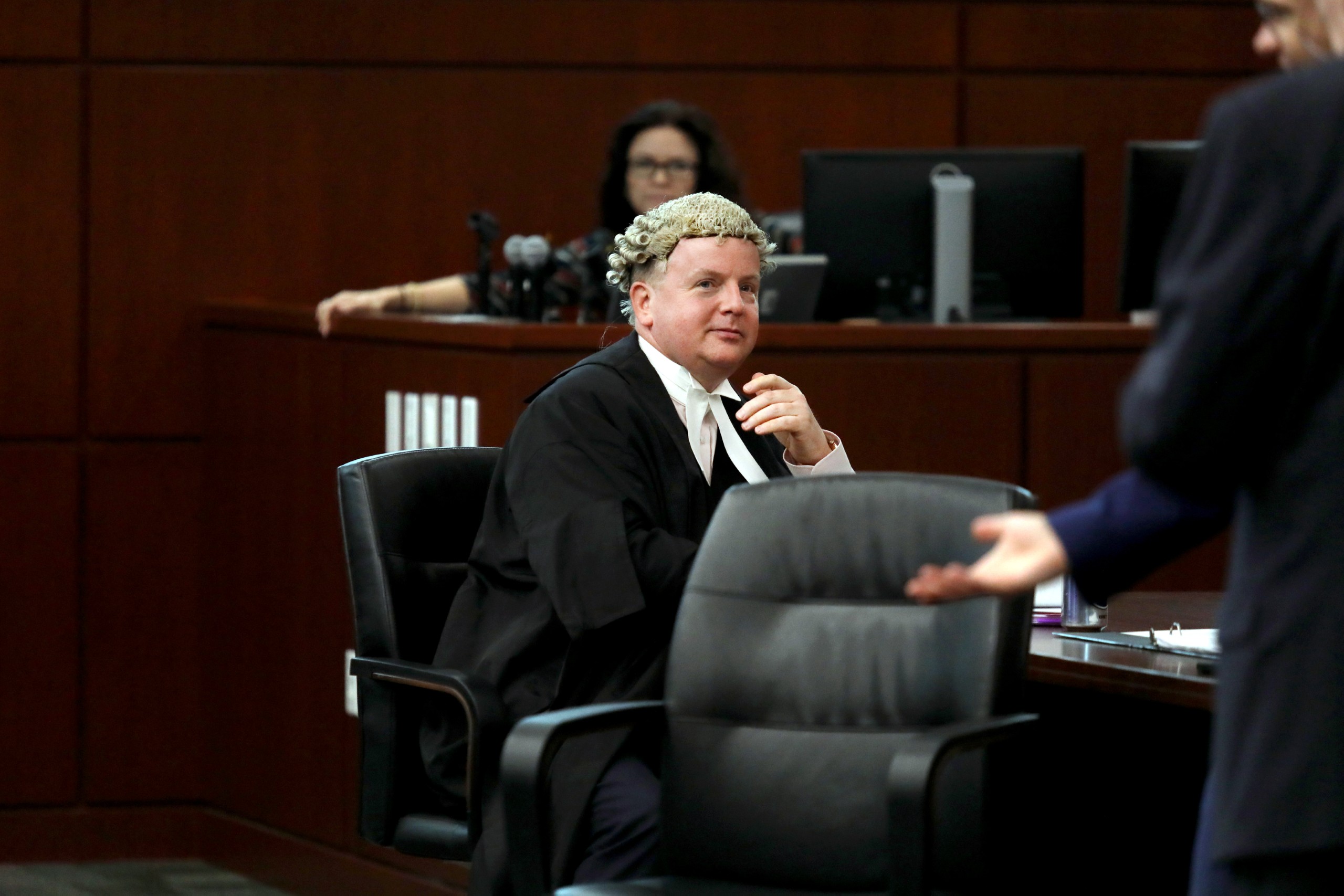 A person wearing a legal gown and wig sits in a courtroom, observing another individual who is gesturing while speaking. There are observers in the background, and the setting includes wooden paneling and computer equipment.