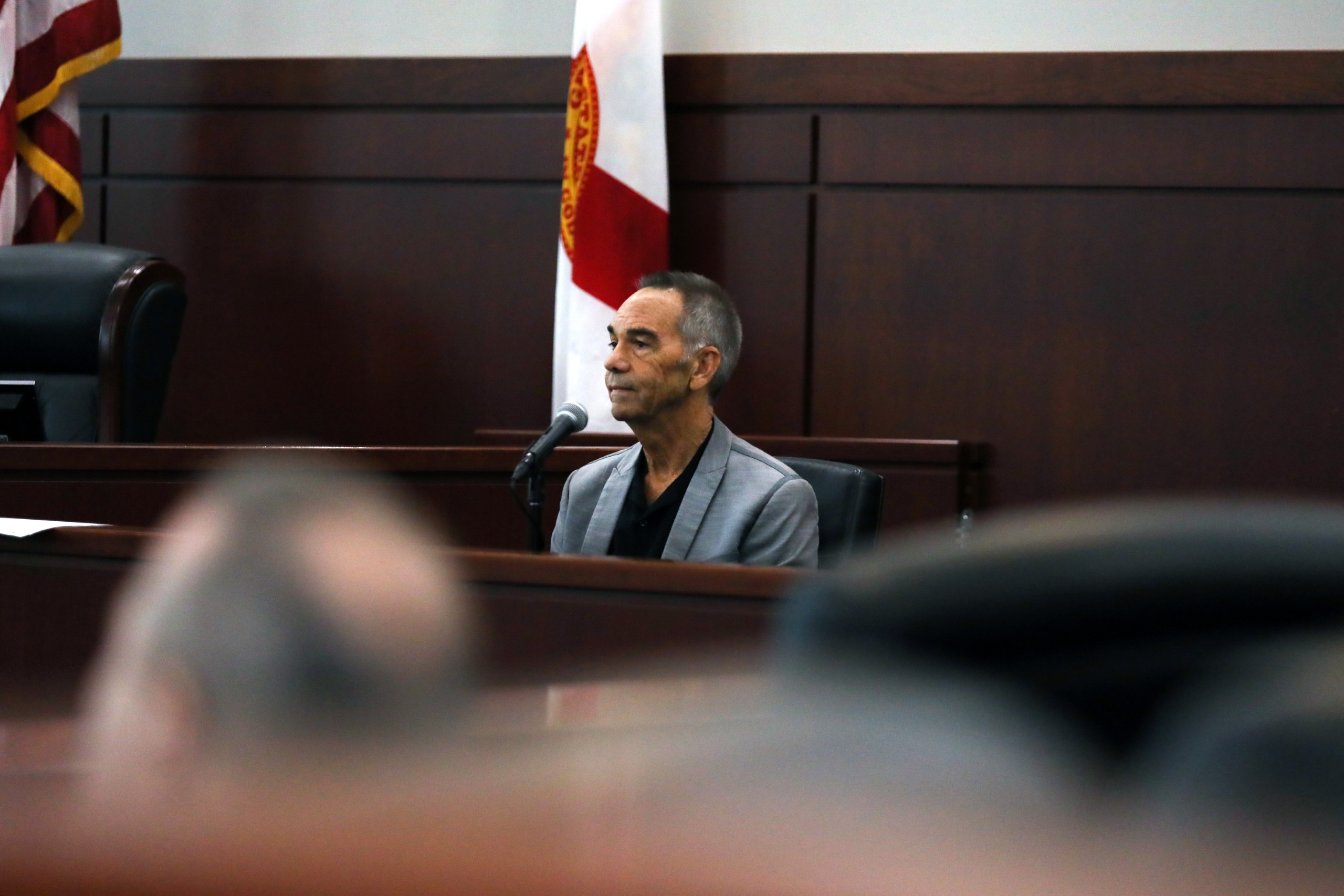 An individual speaks behind a lectern in a courtroom setting, with chairs and flags visible in the background. The focus is on the speaker, who appears engaged in their testimony.
