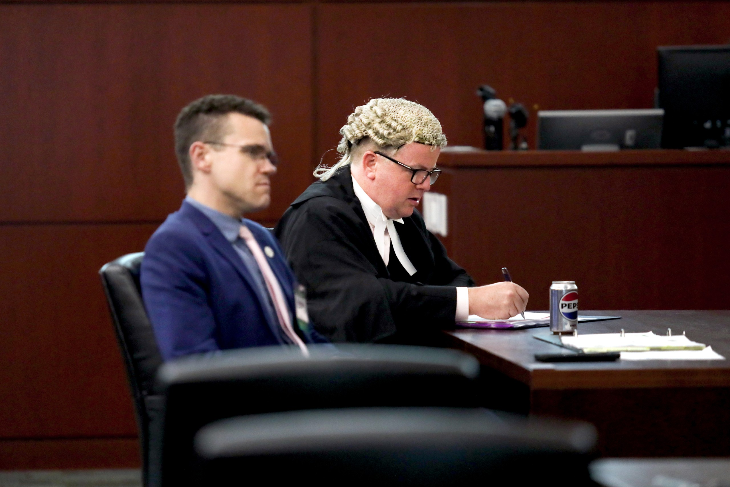 Two individuals sit at a table in a courtroom. One is wearing a wig and writing on paper, while the other is looking toward the front. A can of soda and papers are visible on the table.