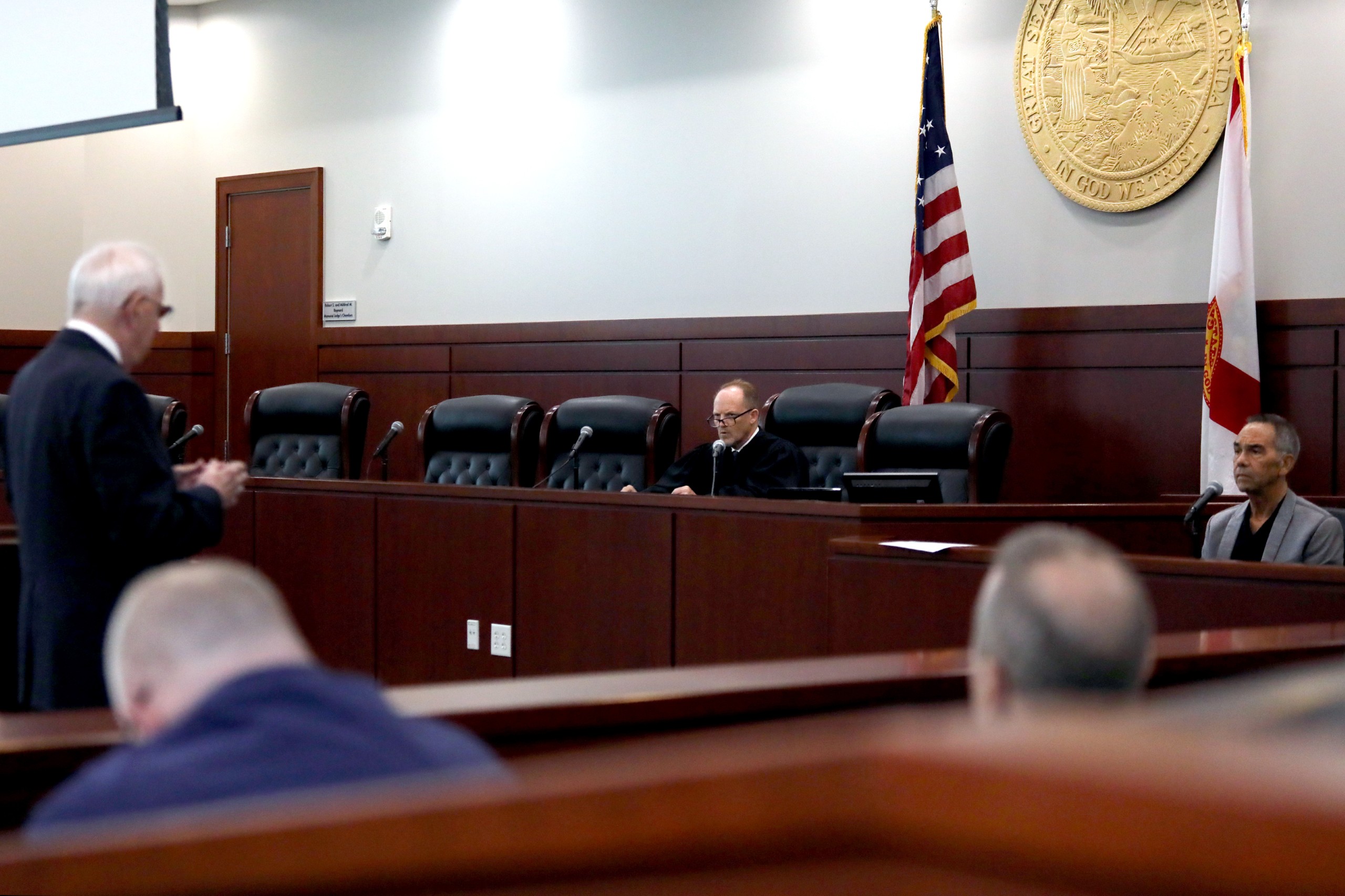 A courtroom scene featuring a person speaking behind a lectern while facing a judge seated at a bench. Flags are positioned in the background, and additional individuals are seated in the gallery.