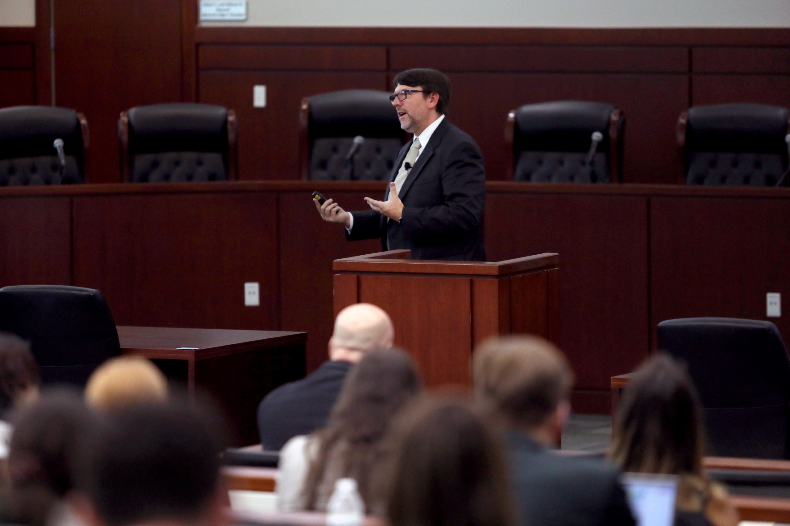A person is speaking behind a lectern to an audience in a formal setting, with several chairs visible in the background. The audience, partially obscured, is engaged in the presentation.