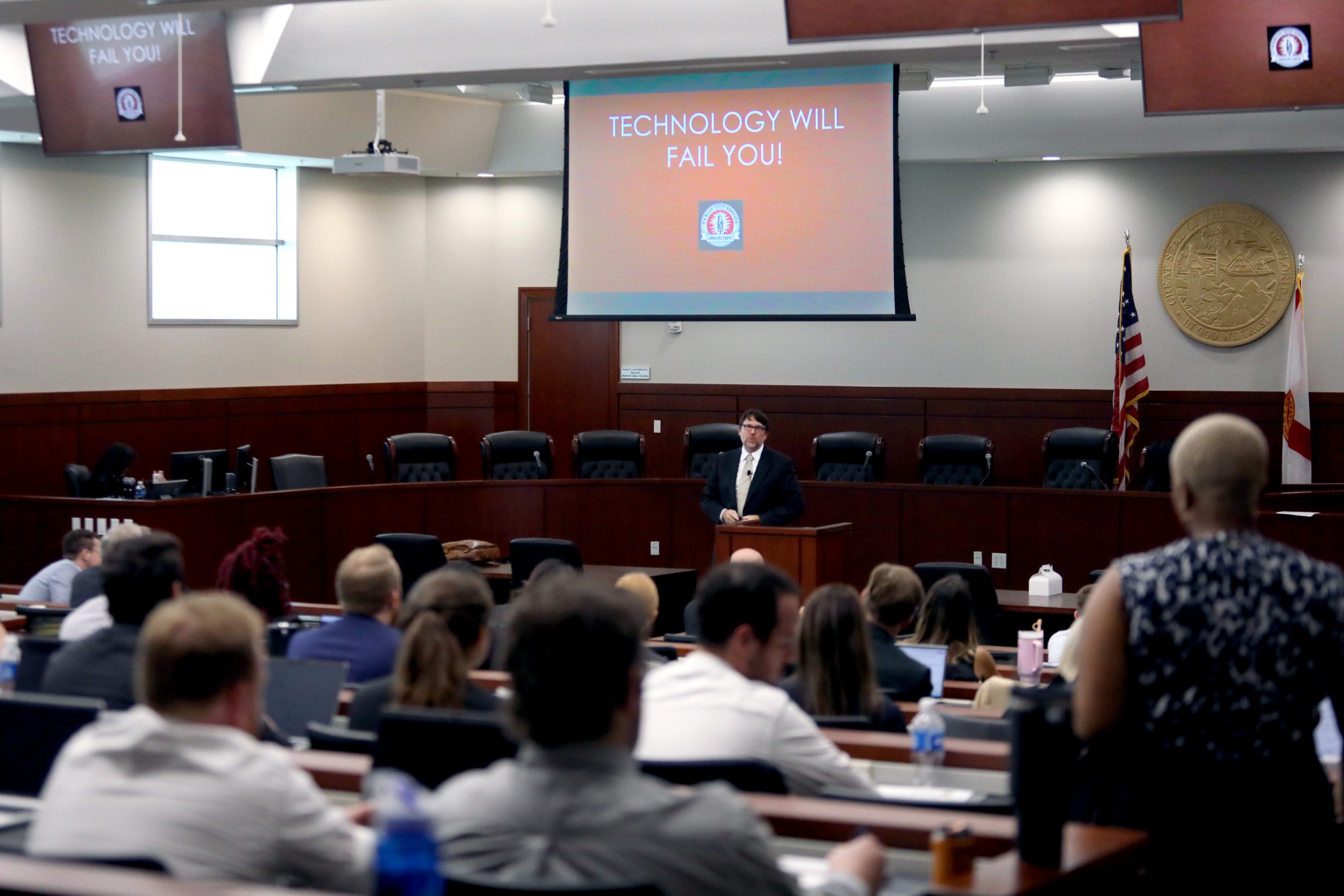 A person stands behind a lectern, speaking to an audience in a classroom setting. A large screen displays the text "TECHNOLOGY WILL FAIL YOU!" Several individuals in the audience are engaged, with some using laptops. The room has a formal appearance, with flags and a seal visible in the background.