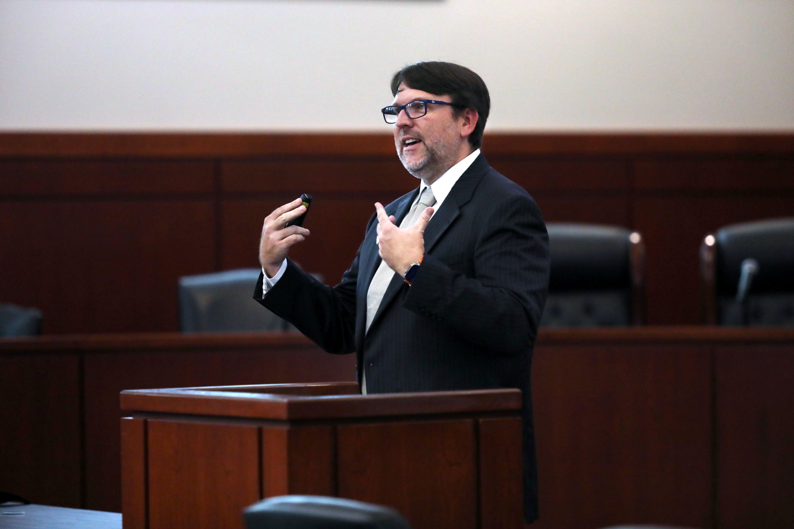 An individual is speaking behind a lectern in a courtroom setting, gesturing with one hand while holding a presentation remote in the other. Rows of empty chairs are visible in the background.