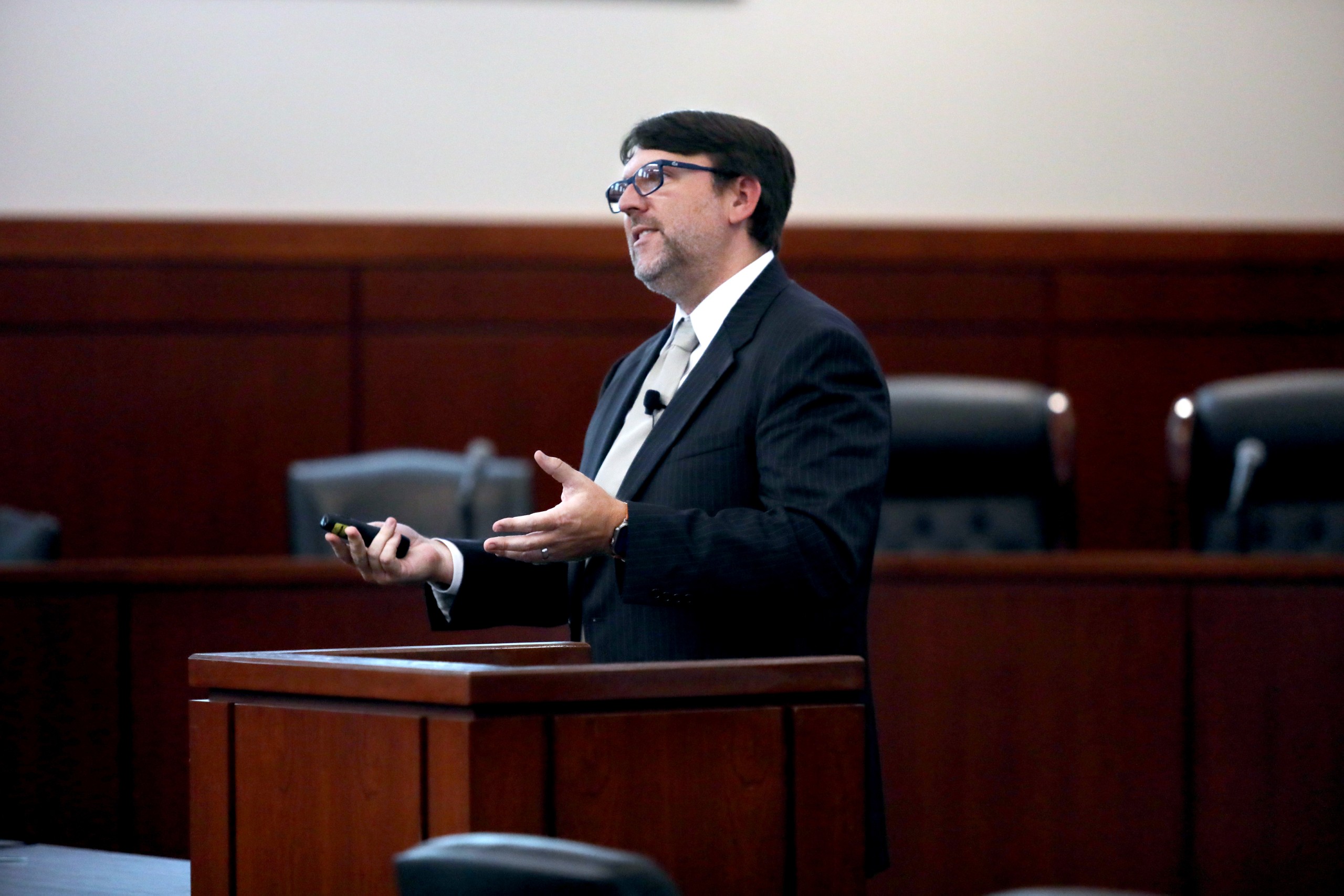A person stands behind a lectern, gesturing while speaking to an audience in a formal setting. The background features empty chairs, suggesting a gathering or presentation event.