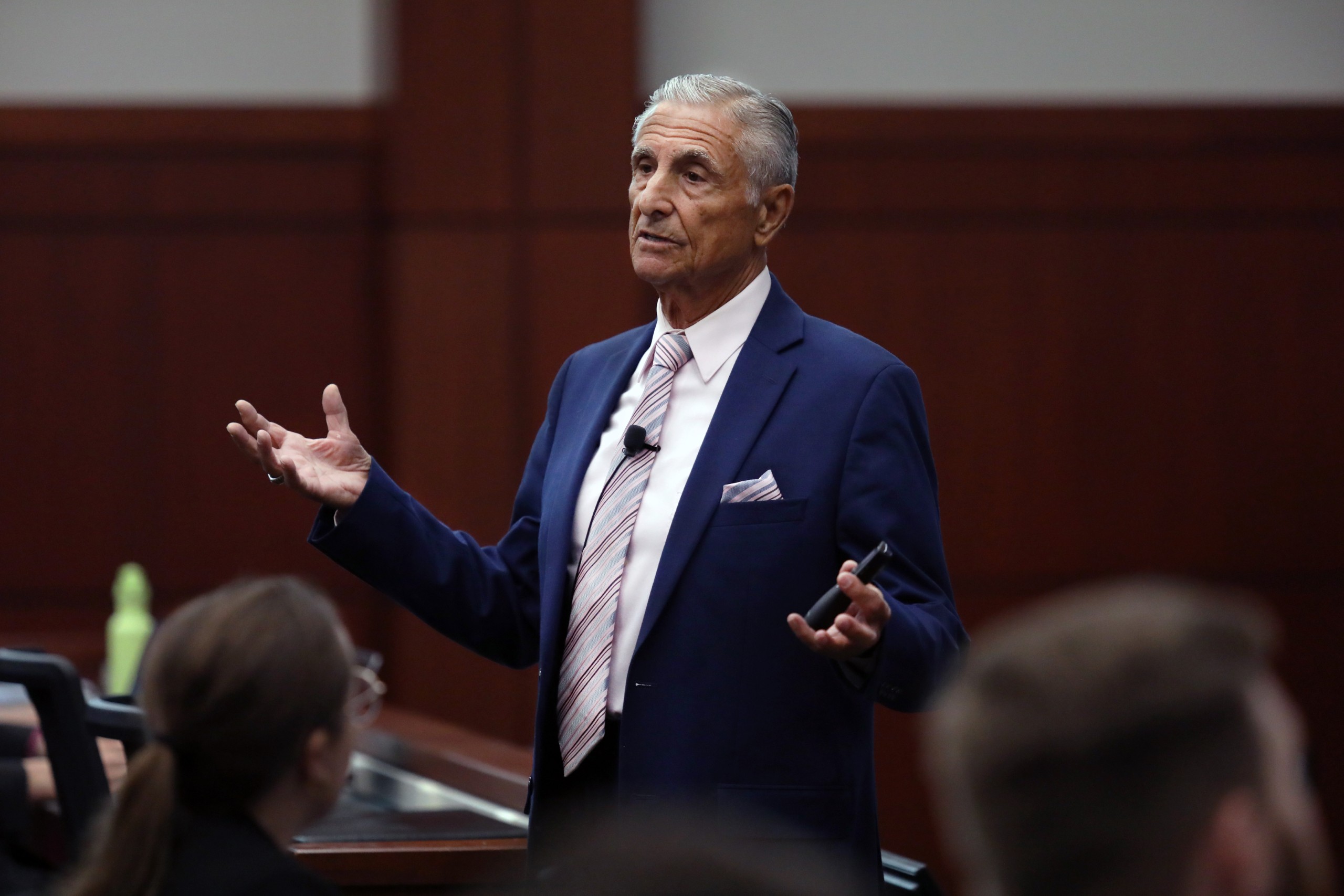 An individual speaks behind a lectern to an audience in a formal setting, gesturing with hands while holding a device.