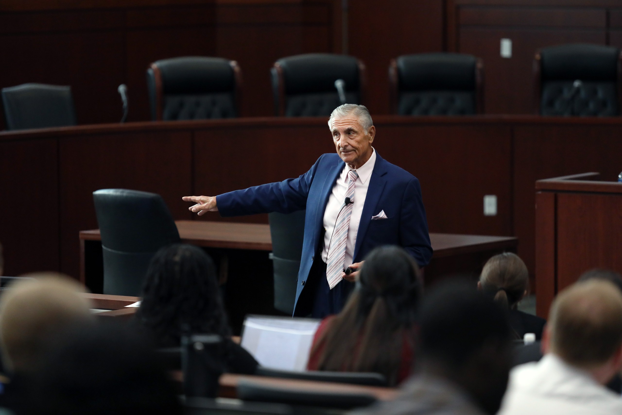 A person is speaking behind a lectern in front of an audience in a courtroom setting. The speaker is gesturing while presenting.