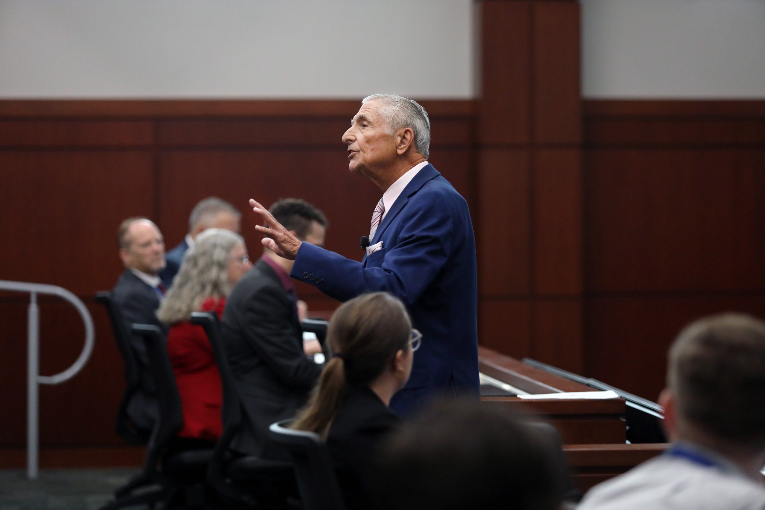 A person stands behind a lectern, speaking to an audience in a courtroom setting. Several individuals are seated in the foreground and background, listening attentively. The room has wooden panels and a formal appearance.
