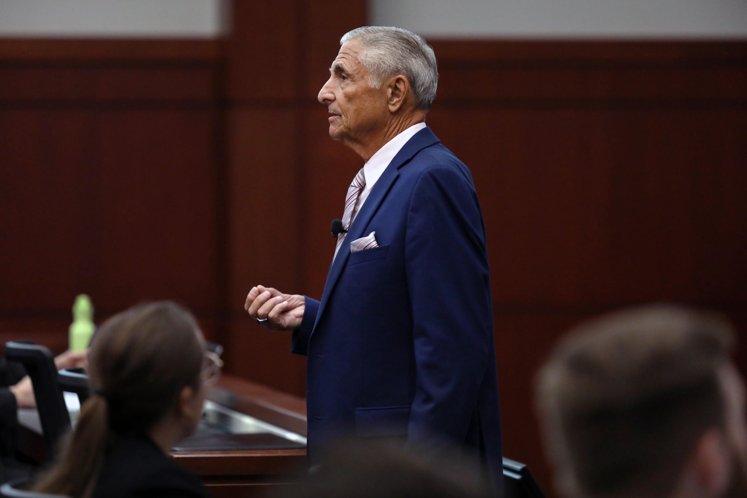 A person stands behind a lectern in a courtroom, gesturing while speaking to an audience. The background features wooden walls and seating.
