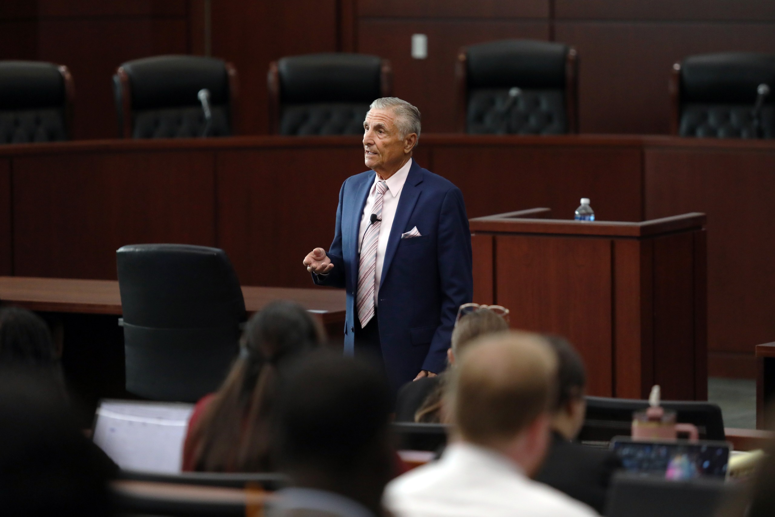 A speaker stands behind a lectern addressing an audience in a formal setting with wooden paneling and seating.