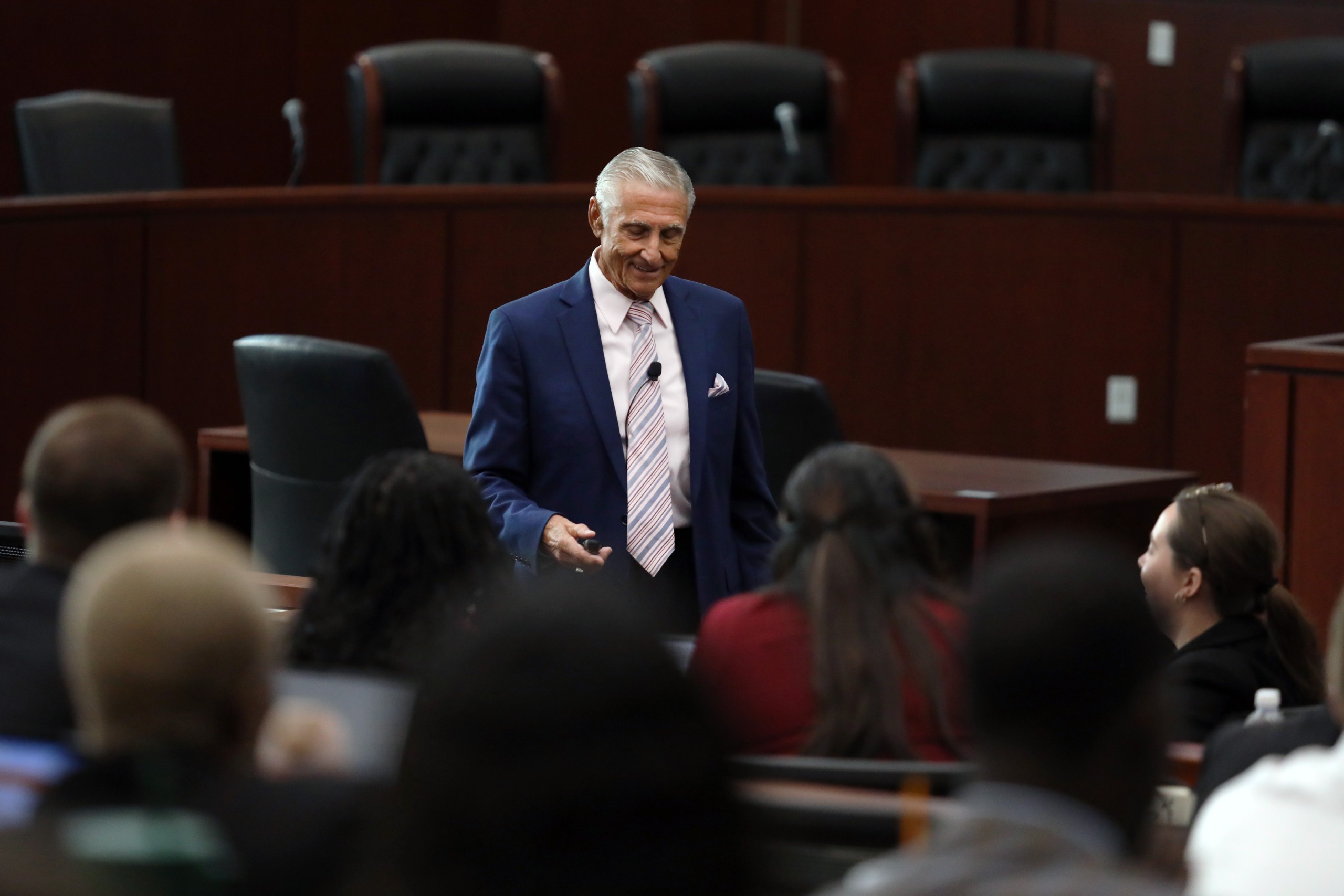A speaker stands behind a lectern addressing an audience in a formal setting with rows of seats. The audience is engaged, facing the speaker.