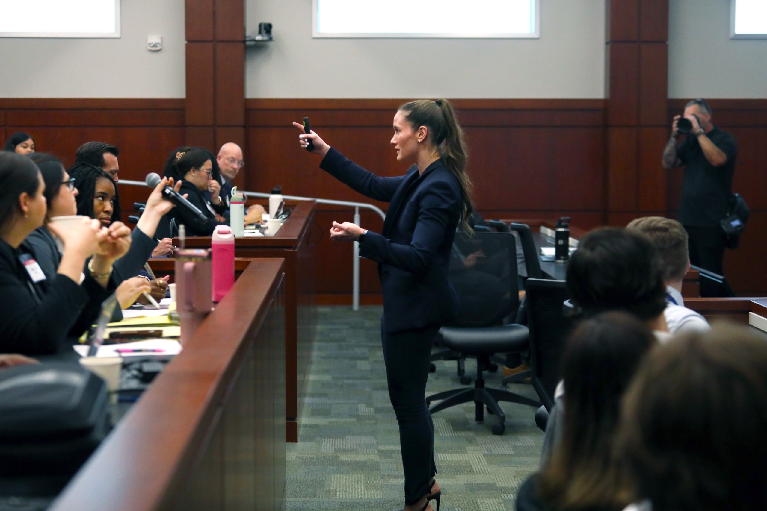 A person stands behind a lectern speaking to an audience in a courtroom setting. The audience, seated in rows, appears engaged and is taking notes.